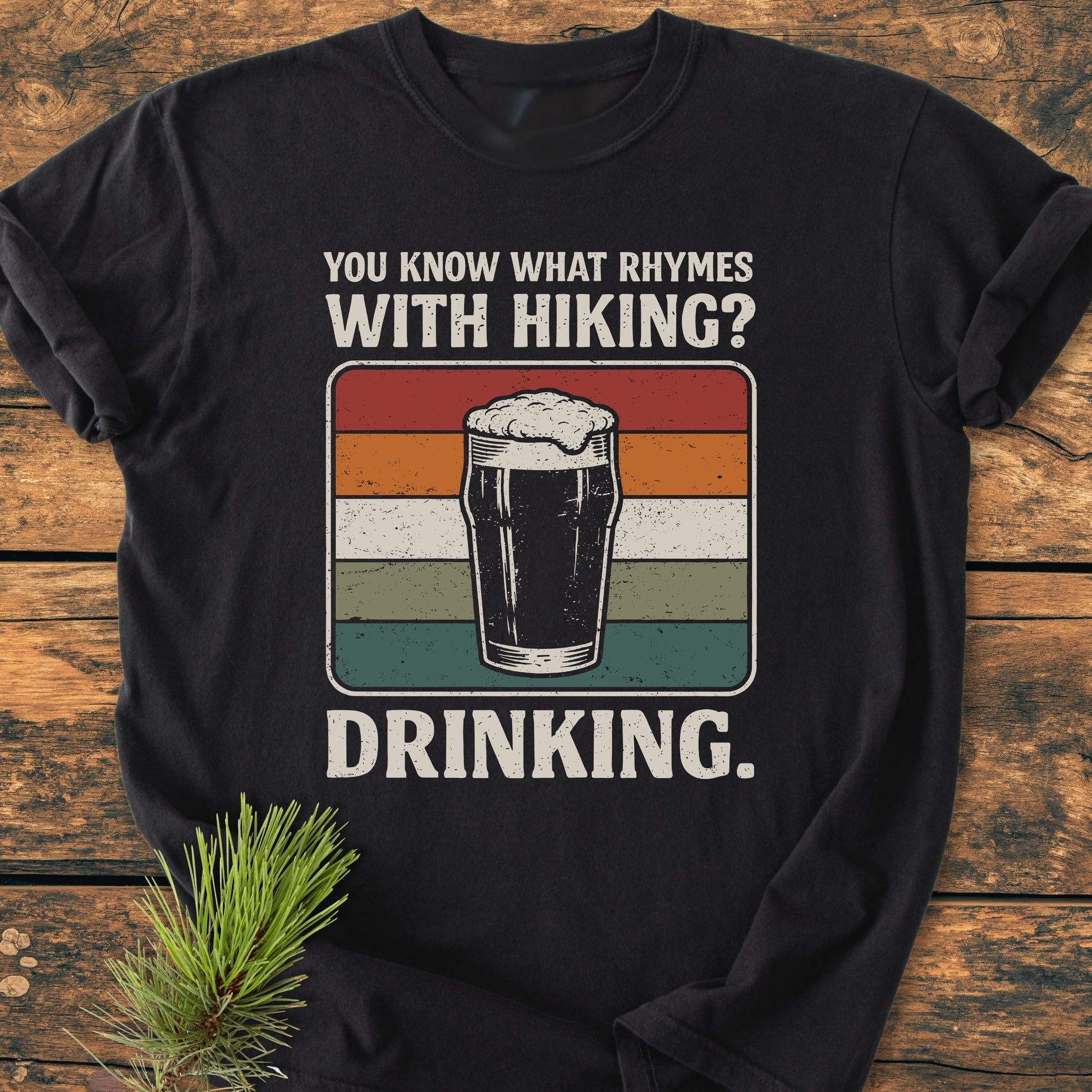 Black T-shirt with retro-styled graphic of a beer pint and text: You know what rhymes with hiking? DRINKING. Laid on wood with pine sprig; embodies What Rhymes With Hiking? - Vintage Wash Tee.
