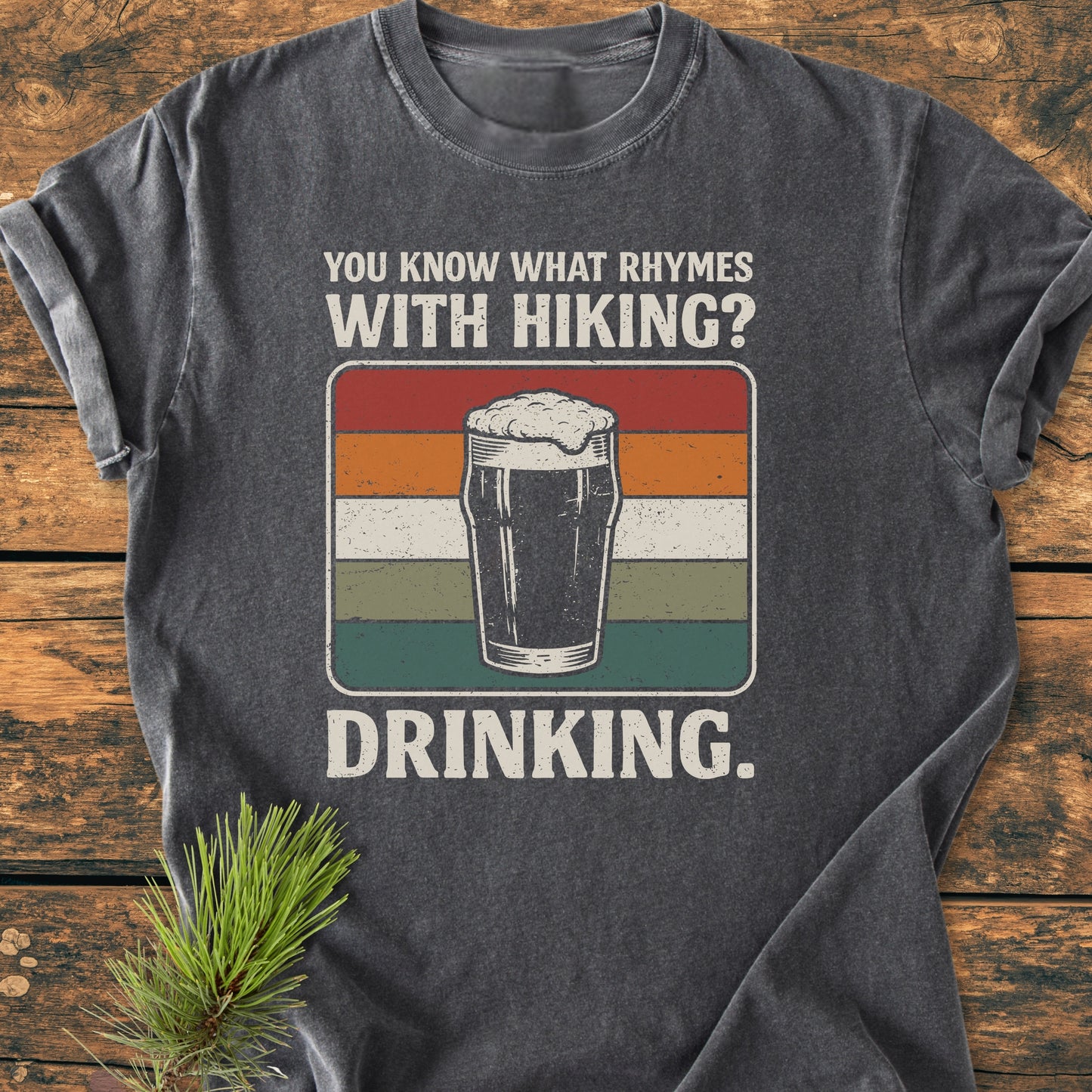 Dark-gray tee with retro pint graphic, You Know What Rhymes With Hiking? Drinking., laid on wood with pine branch; embodies trail humor, perfect for adventure-loving hikers and beer enthusiasts.