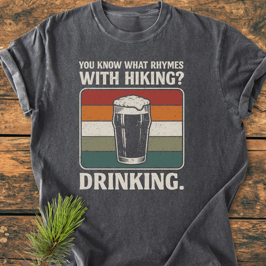 Dark-gray tee with retro pint graphic, You Know What Rhymes With Hiking? Drinking., laid on wood with pine branch; embodies trail humor, perfect for adventure-loving hikers and beer enthusiasts.
