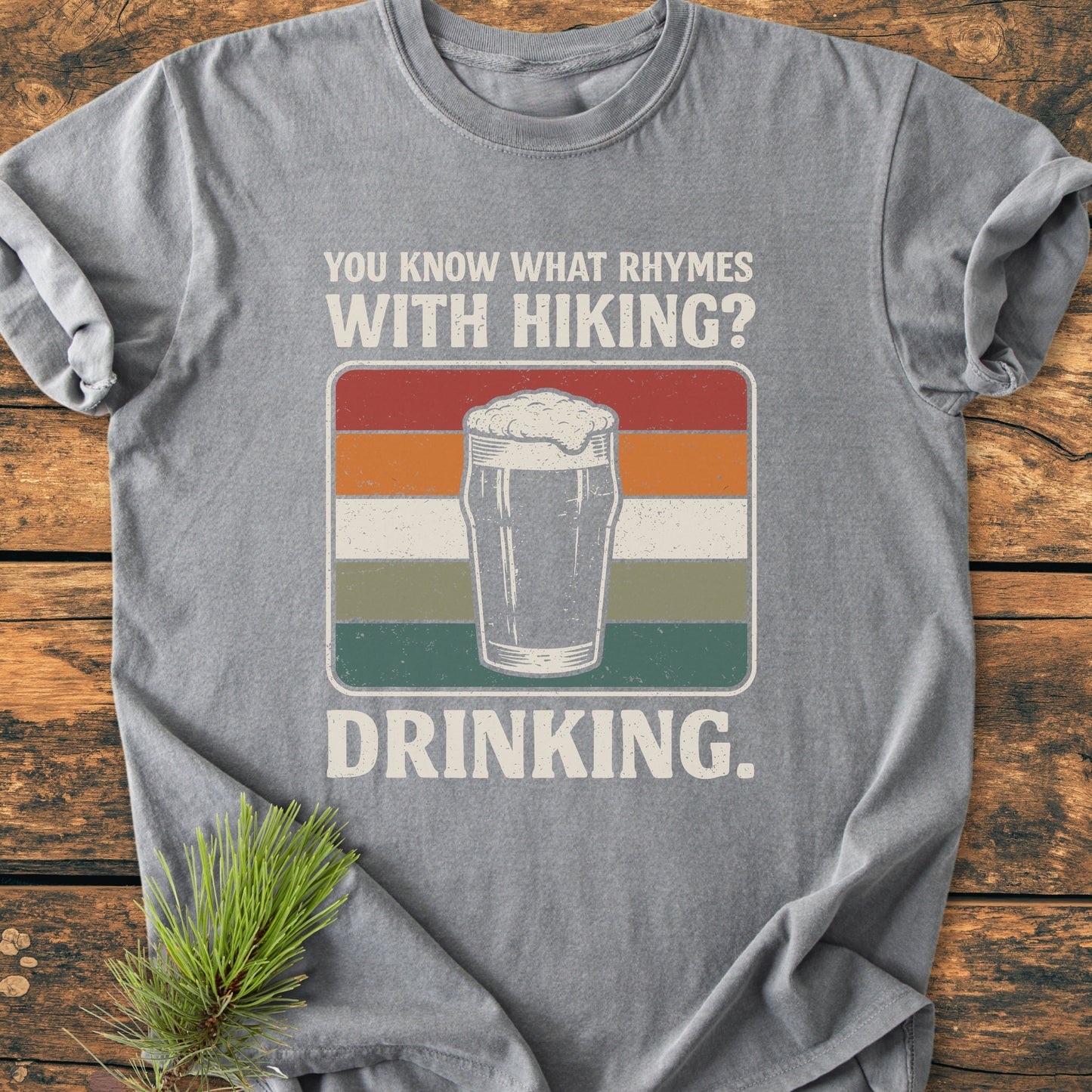 What Rhymes With Hiking? - Vintage Wash Tee featuring a retro pint glass design and humorous slogan, flat-lay on wood with pine sprig, ideal for trail-loving beer enthusiasts.