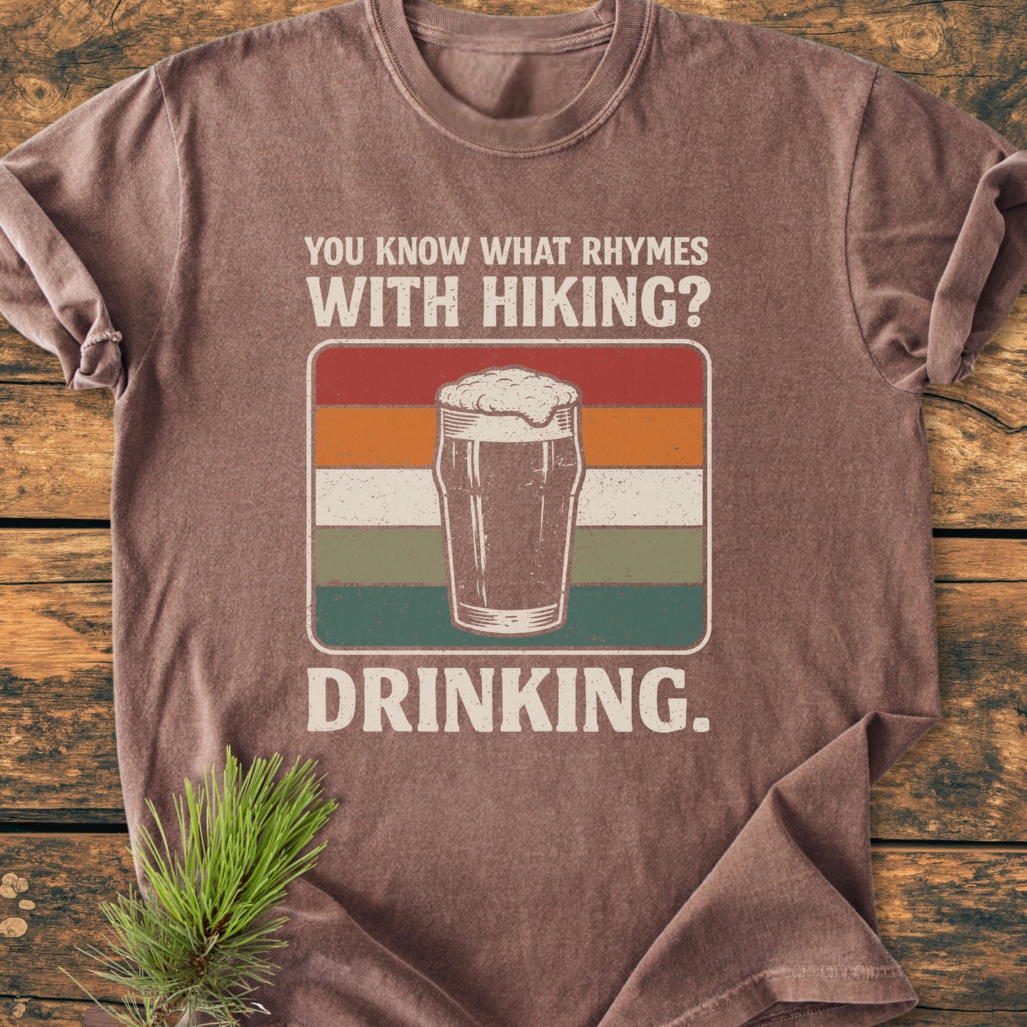 What Rhymes With Hiking? - Vintage Wash Tee featuring a retro frothy pint graphic with humorous text, laid on wood with pine sprig, epitomizes trail humor and post-hike relaxation.