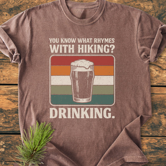 What Rhymes With Hiking? - Vintage Wash Tee featuring a retro frothy pint graphic with humorous text, laid on wood with pine sprig, epitomizes trail humor and post-hike relaxation.