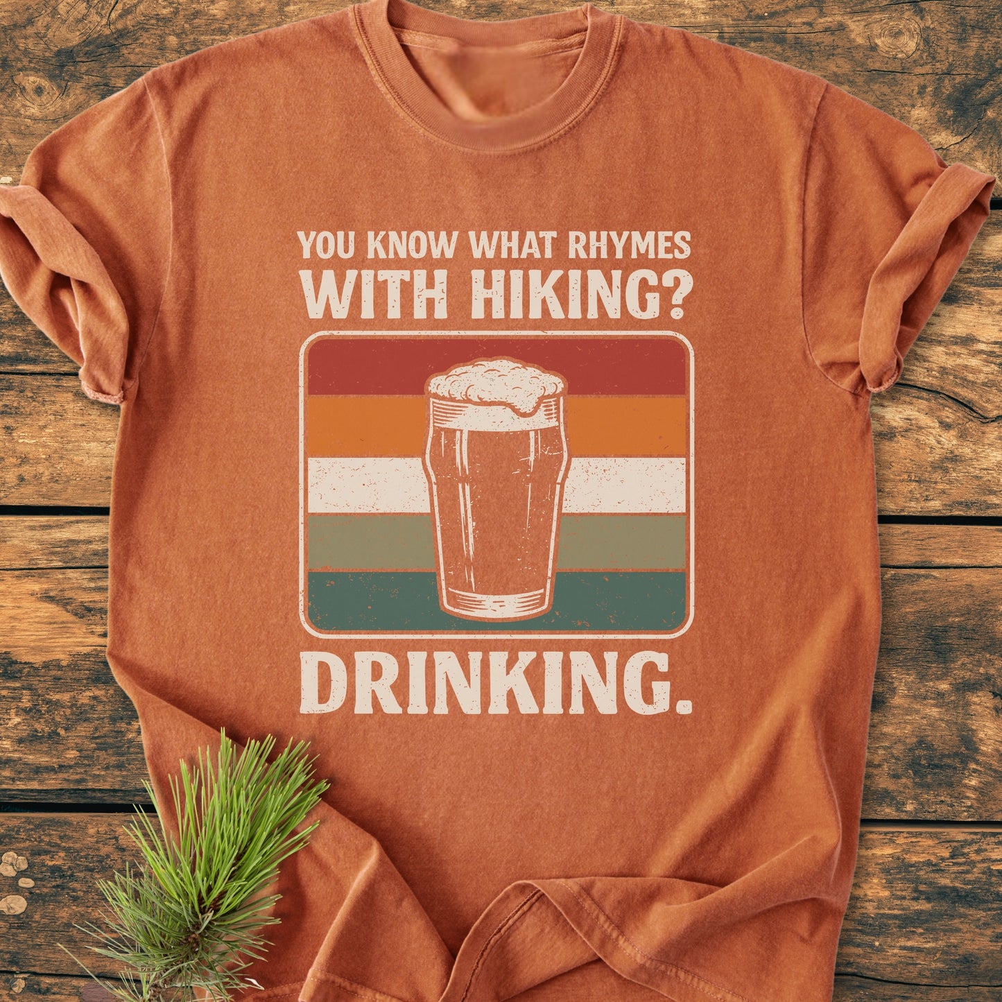 Rust-orange What Rhymes With Hiking? - Vintage Wash Tee featuring a frothy pint glass drawing and slogan, laid on a wooden surface with a pine sprig, highlighting outdoor humor and comfort.