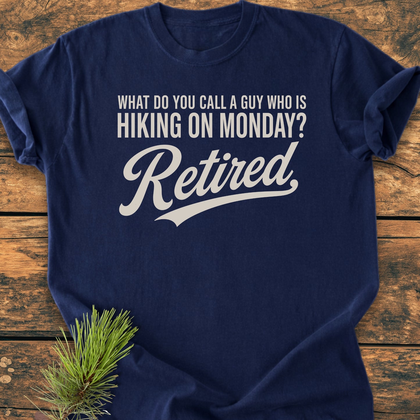 Hiking On Monday - Vintage Wash Tee features a navy-blue color with cream lettering of a humorous retirement joke, laid on wood with a pine sprig, highlighting its relaxed, comfortable style.