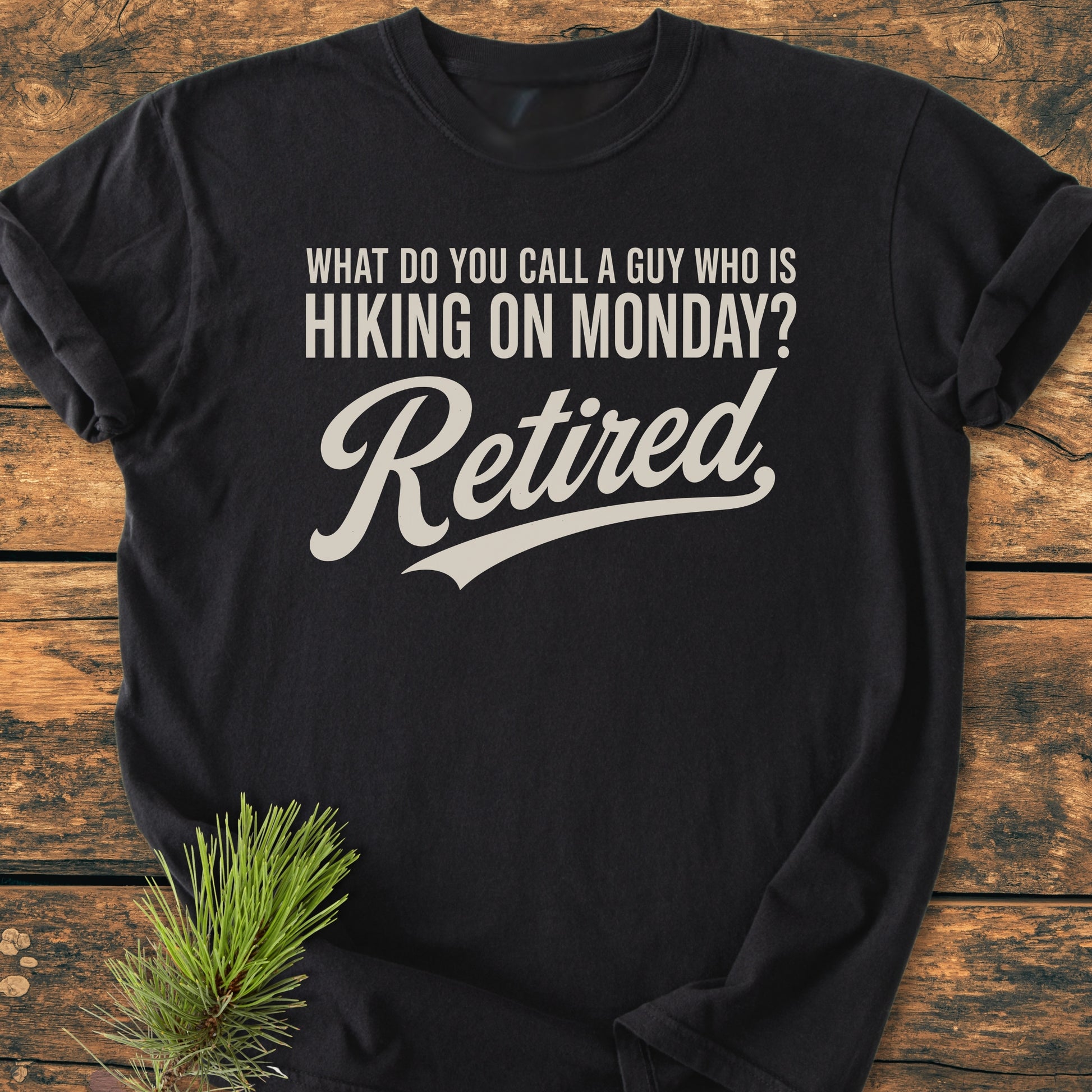 Hiking On Monday - Vintage Wash Tee displayed on a wooden surface with a pine sprig, featuring a humorous retirement-themed hiking joke in bold white lettering. Perfect for outdoor-loving retirees.