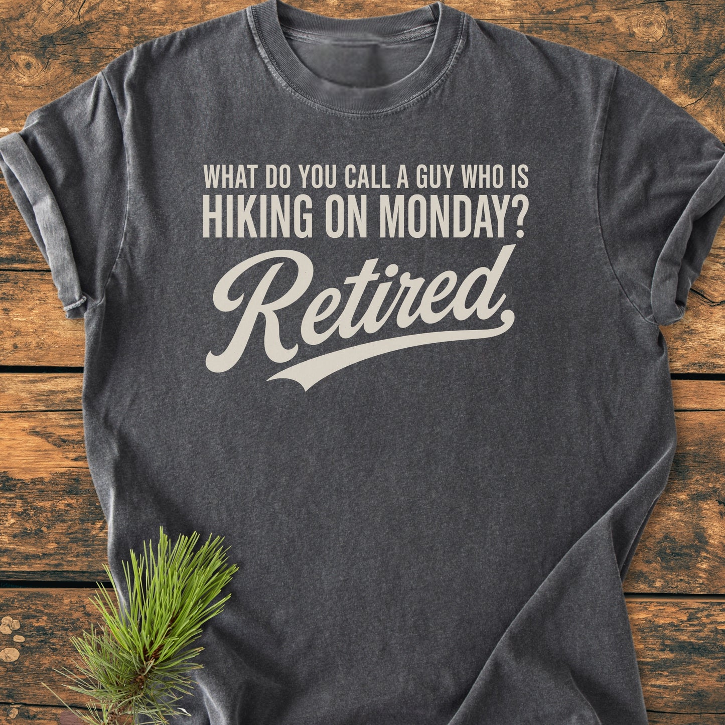 Hiking On Monday - Vintage Wash Tee: Charcoal-gray T-shirt with cream-colored humorous hiking text, styled with a pine branch, laid on wood, embodying outdoor adventure and relaxed retirement spirit.