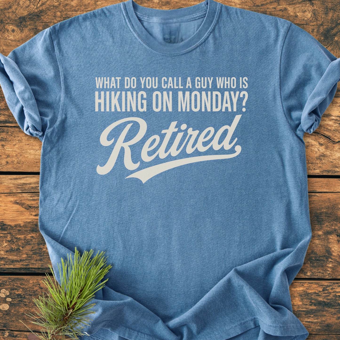 Heather-blue T-shirt with “Hiking On Monday - Vintage Wash Tee” printed in off-white. Features a playful retirement joke, laid flat on a wooden surface, showcasing its relaxed fit and vintage appeal.