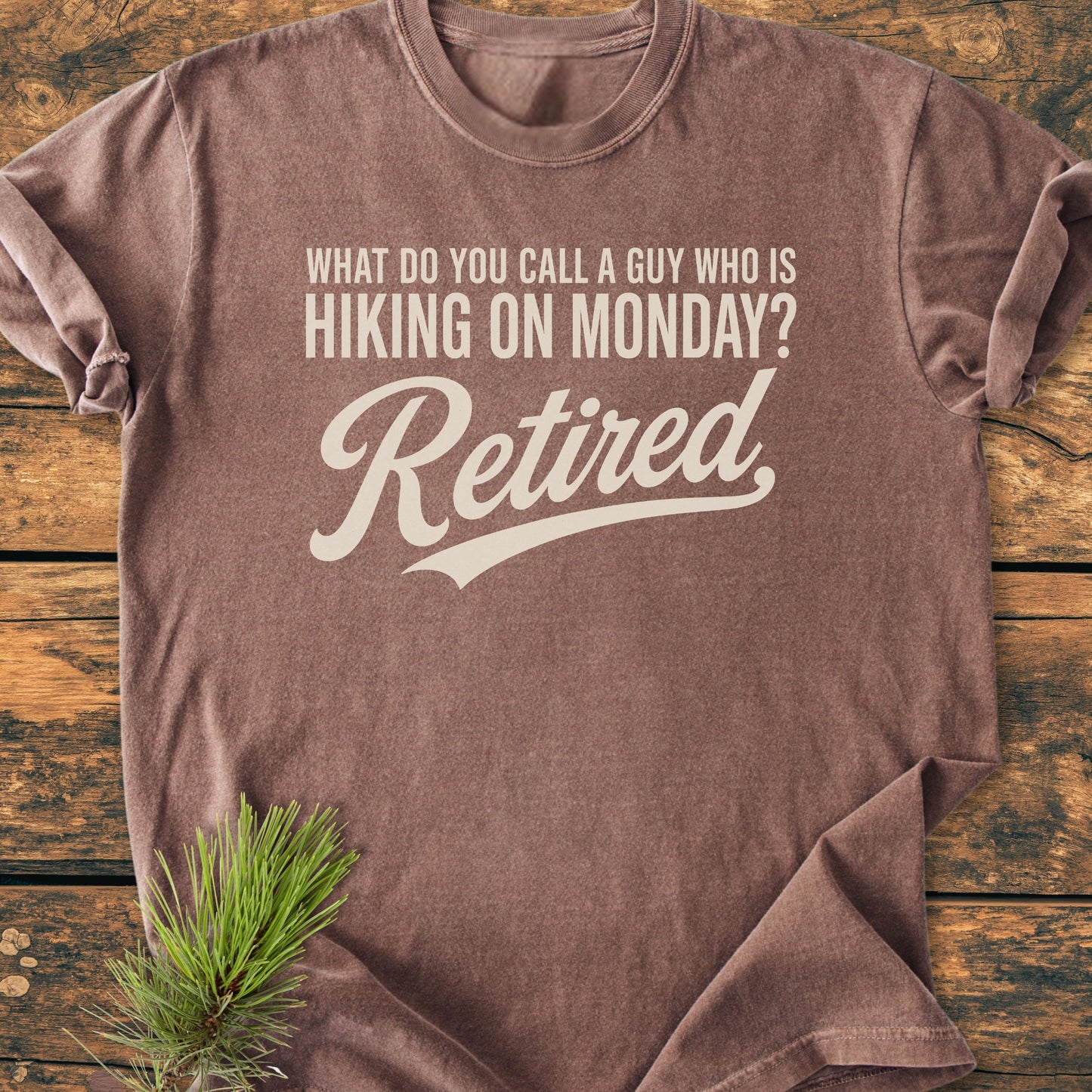 Hiking On Monday - Vintage Wash Tee, featuring a cream joke print on a brown short-sleeve, laid on rustic wood with a pine sprig. Perfect for retired outdoor enthusiasts.