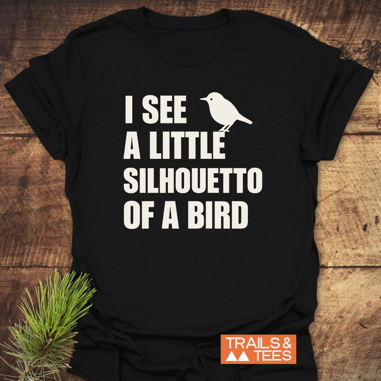 Silhouetto Of A Bird Funny Softstyle T-Shirt on wood, features “I SEE A LITTLE SILHOUETTO OF A BIRD” text with bird silhouette. Displays TRAILS & TEES logo, highlighting lightweight, comfortable design.