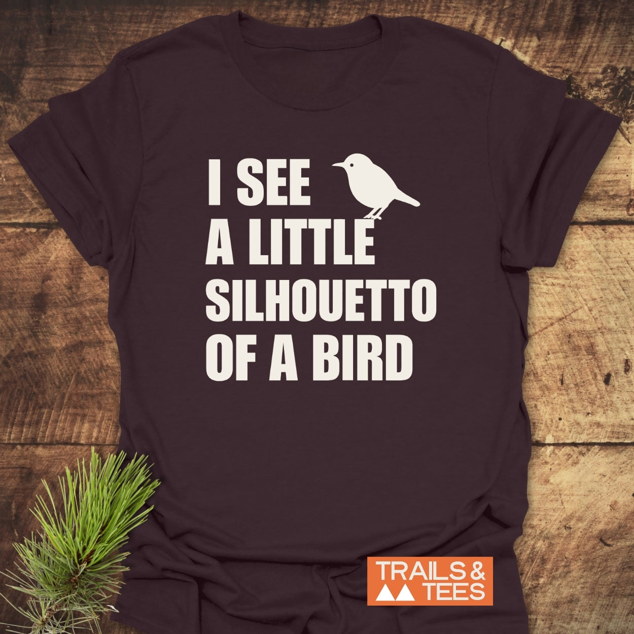 Silhouetto Of A Bird Funny Softstyle T-Shirt on wooden surface, features bold text and bird graphic, embodies everyday comfort with lightweight, smooth fabric. TRAILS & TEES logo in corner.