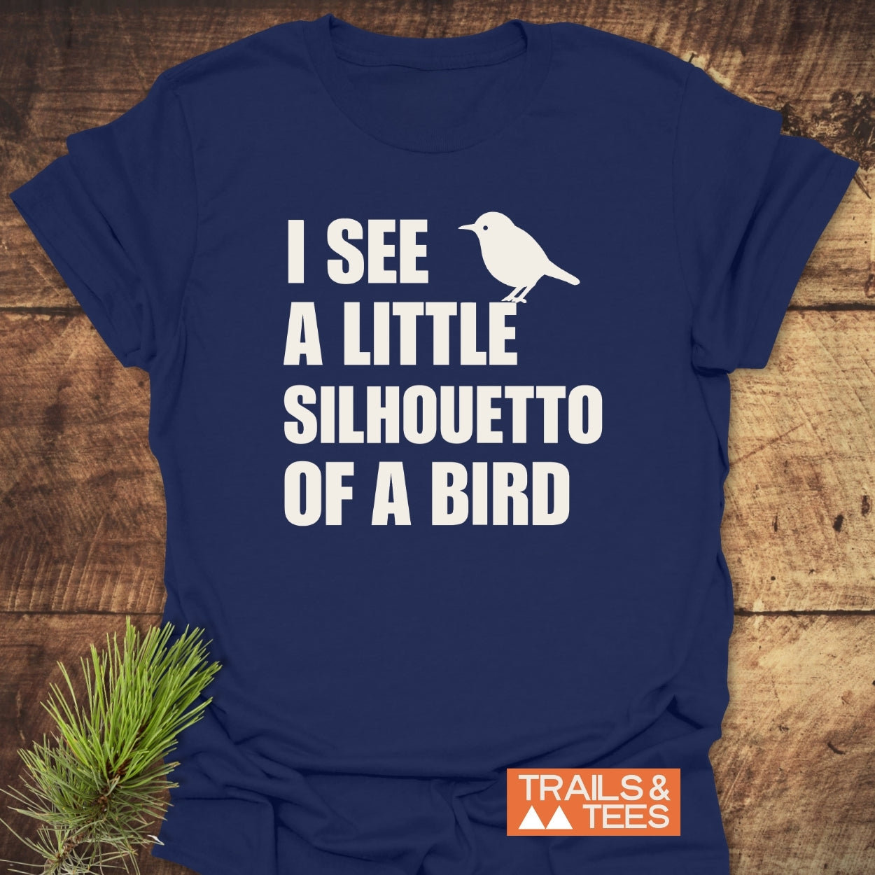 Silhouetto Of A Bird Funny Softstyle T-Shirt featuring a white bird silhouette and text on a navy-blue tee, showcasing a Trails & Tees logo, laid on a wooden surface.