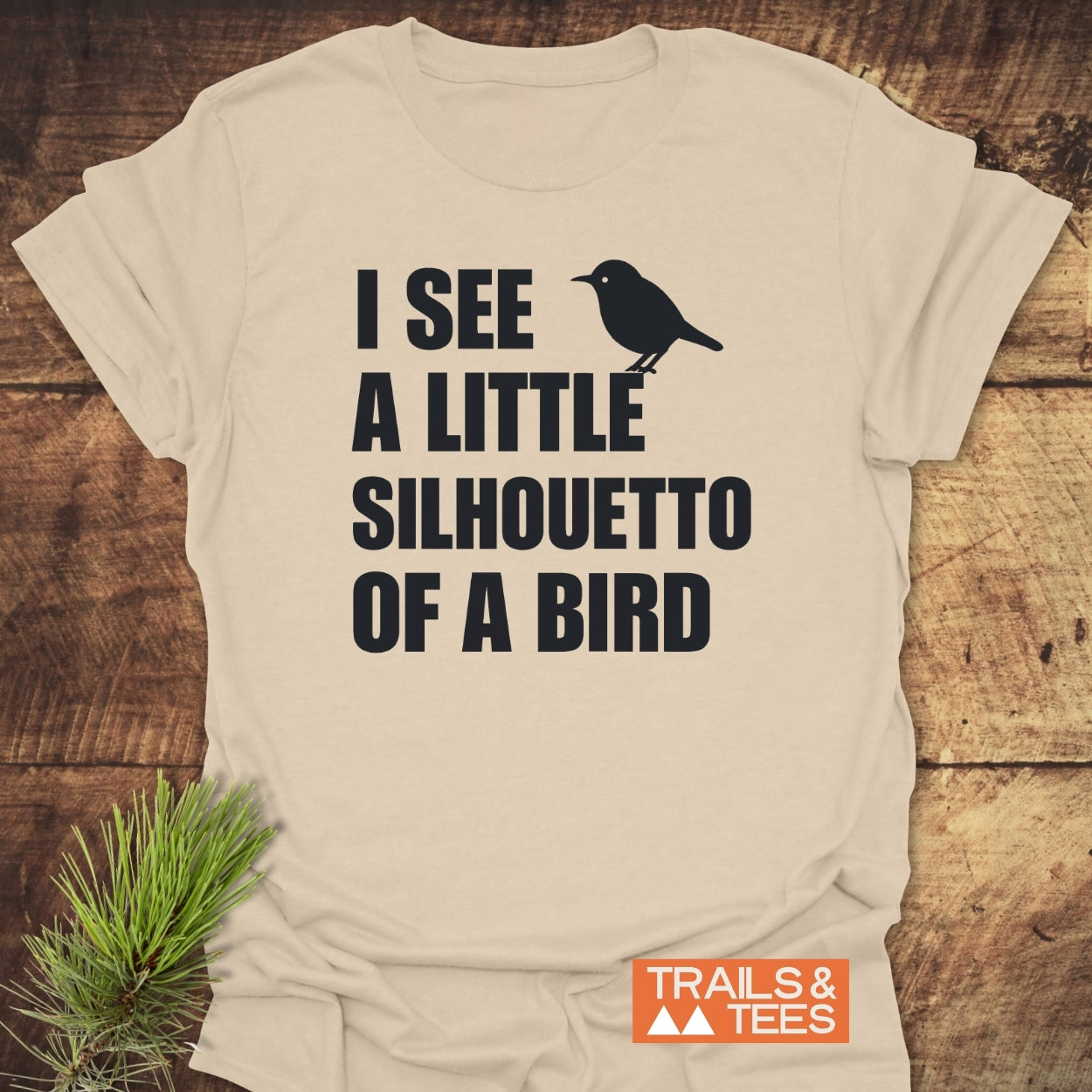 Silhouetto Of A Bird Funny Softstyle T-Shirt featuring bold black text and a small bird silhouette, laid on wood with a pine needle sprig, embodies everyday comfort and outdoor-inspired design.