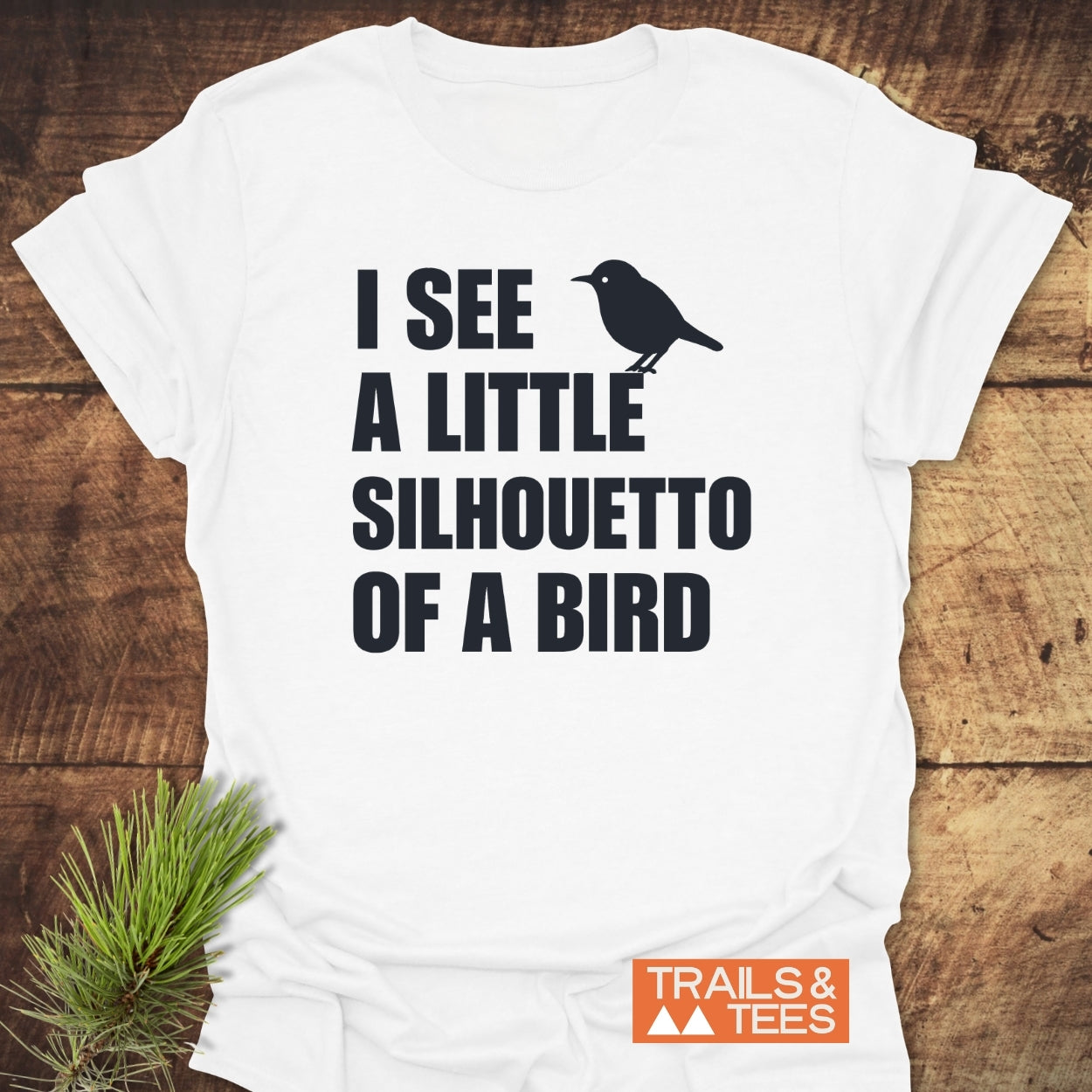 Silhouetto Of A Bird Funny Softstyle T-Shirt on wooden surface with pine sprig, features bold phrase with bird silhouette. Includes Trails & Tees logo, highlighting its lightweight, smooth, everyday-comfort design.