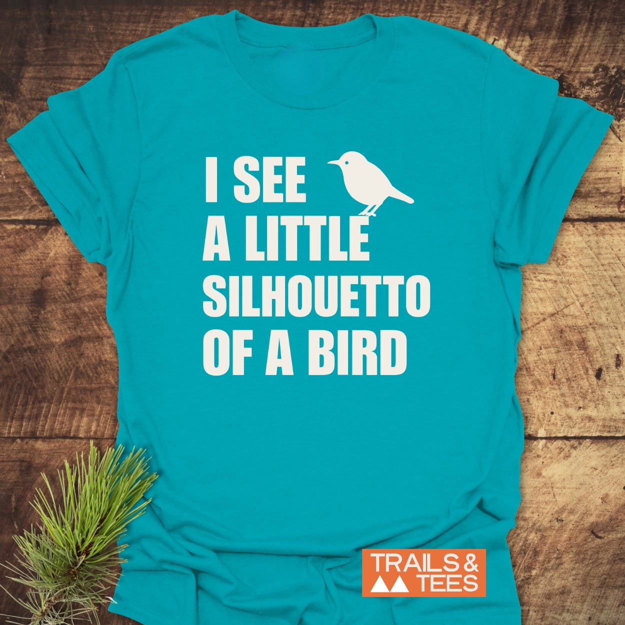 Silhouetto Of A Bird Funny Softstyle T-Shirt displayed on a wooden surface, featuring a white bird silhouette and playful text. Trails & Tees logo and pine sprig accentuate the shirt's outdoor-inspired design.