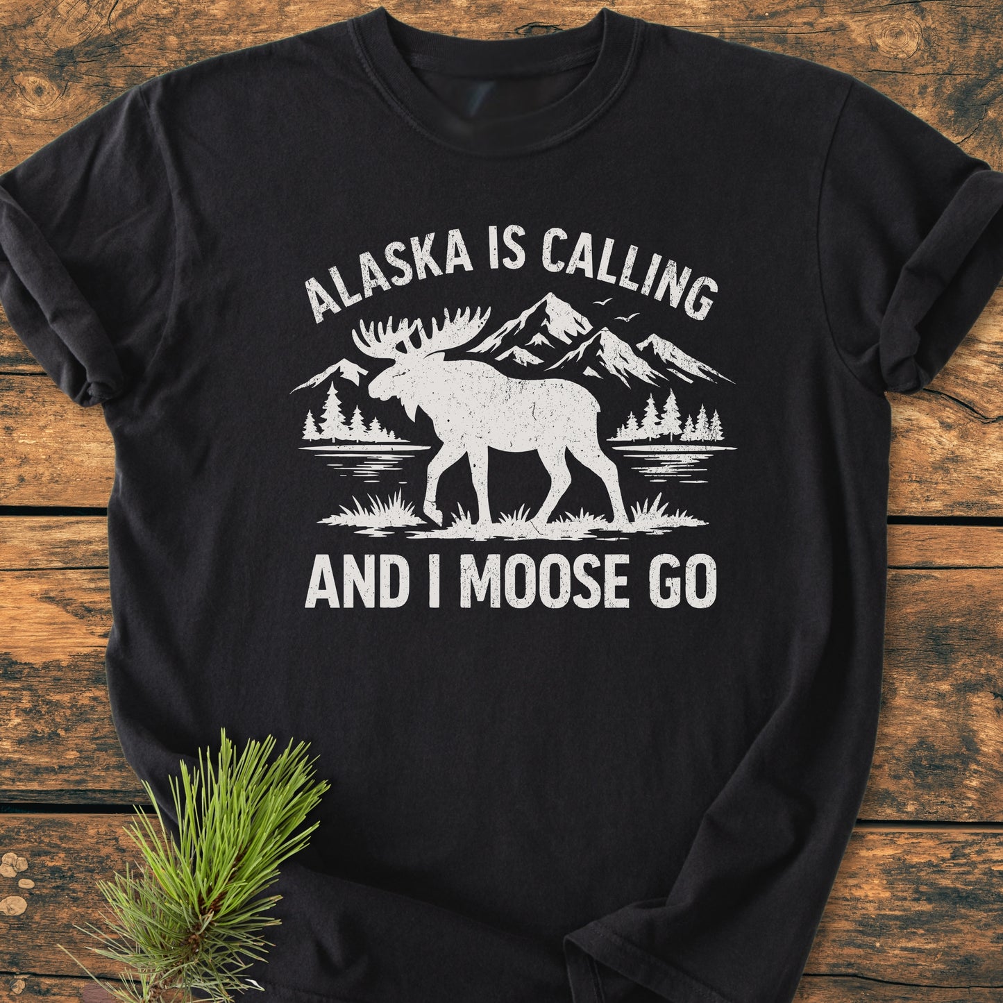 Alaska Is Calling - Vintage Wash Tee features a moose graphic with mountains, trees, and lake on a black T-shirt, styled with a pine branch, highlighting its soft, broken-in design.
