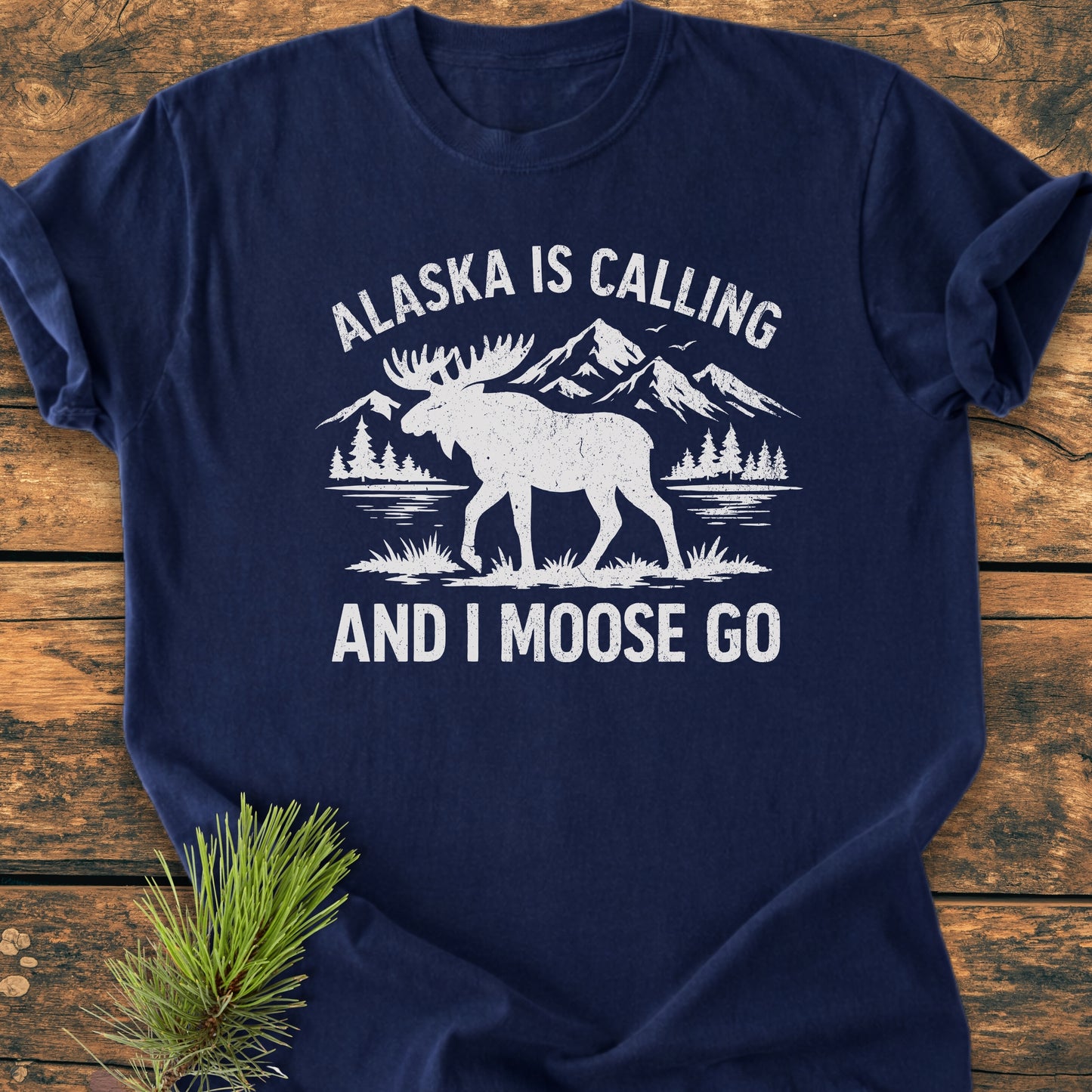 Alaska Is Calling - Vintage Wash Tee, featuring a moose graphic with pine trees and mountains, showcases a relaxed fit on a wooden surface, embodying a soft, lived-in feel.