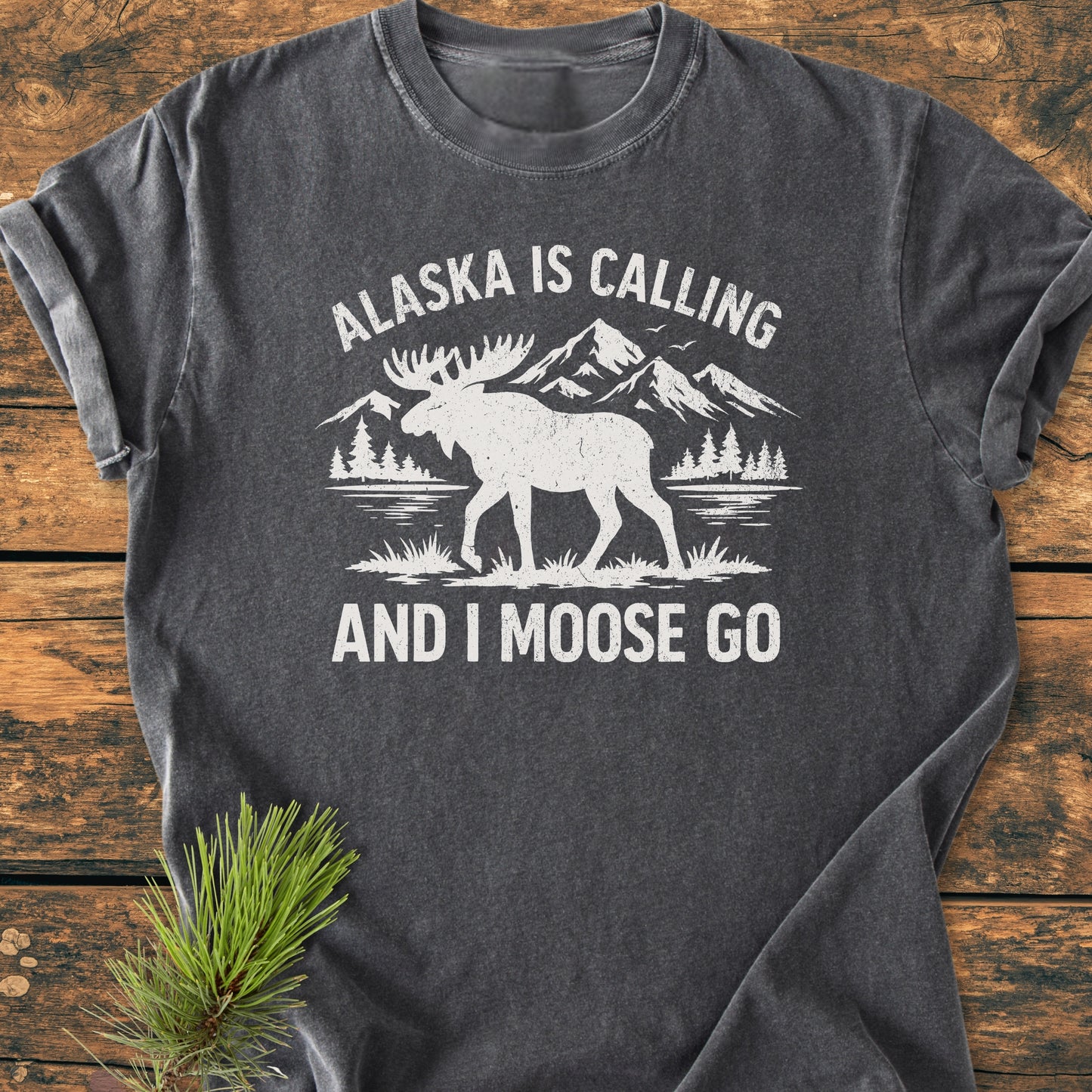 Alaska Is Calling - Vintage Wash Tee featuring a moose graphic with mountains and trees on a charcoal-gray backdrop, laid on wood with pine sprig, perfect for outdoor enthusiasts.