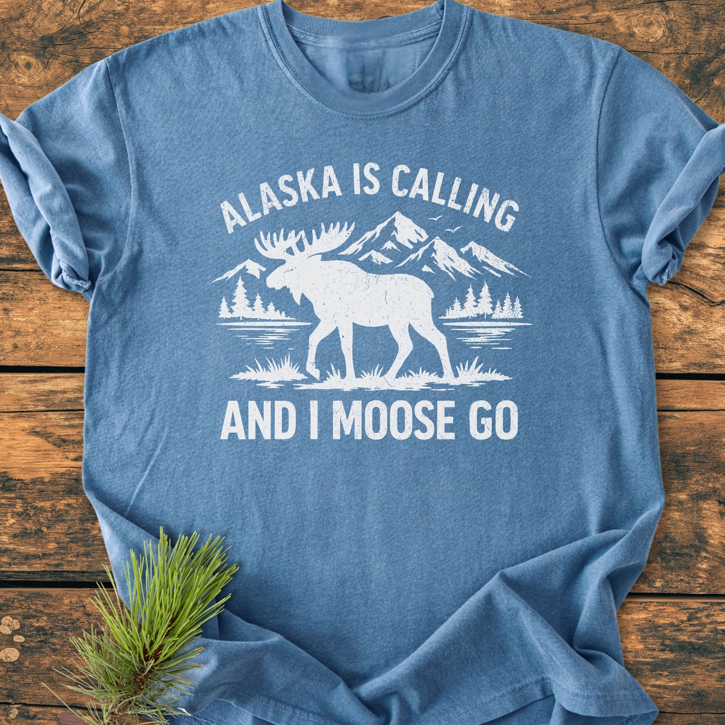 Heather-blue Alaska Is Calling - Vintage Wash Tee features a moose silhouette against a mountain lakescape with a pun slogan. Laid flat, showcasing its relaxed fit and vintage character, beside a pine sprig.