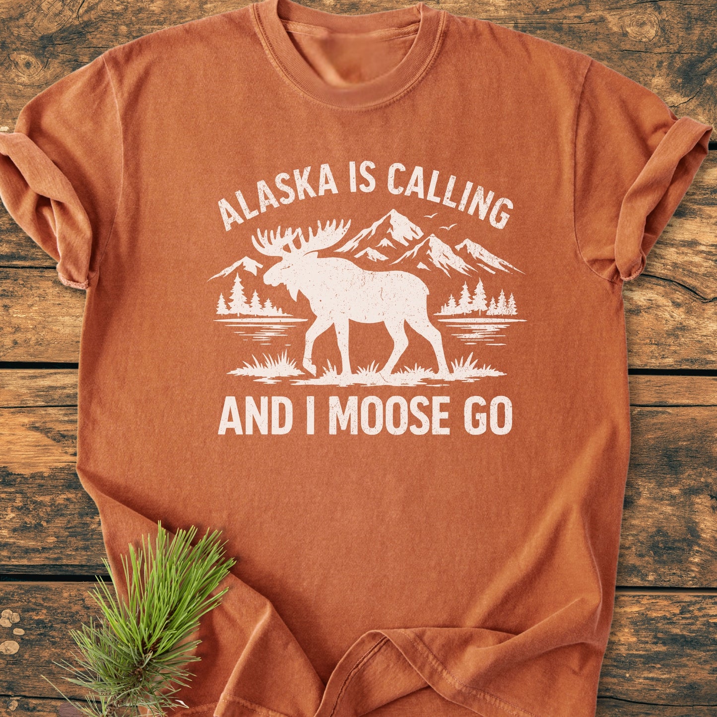 Alaska Is Calling - Vintage Wash Tee features a white moose graphic with mountains and forest on a rust-orange shirt, styled with a pine sprig, showcasing a relaxed fit and vintage wash.