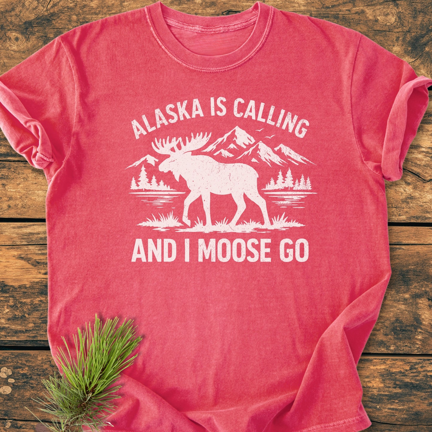 Alaska Is Calling - Vintage Wash Tee: Heather-red T-shirt featuring a white wilderness scene with moose silhouette, laid on wood. Text reads “ALASKA IS CALLING” and “AND I MOOSE GO.”