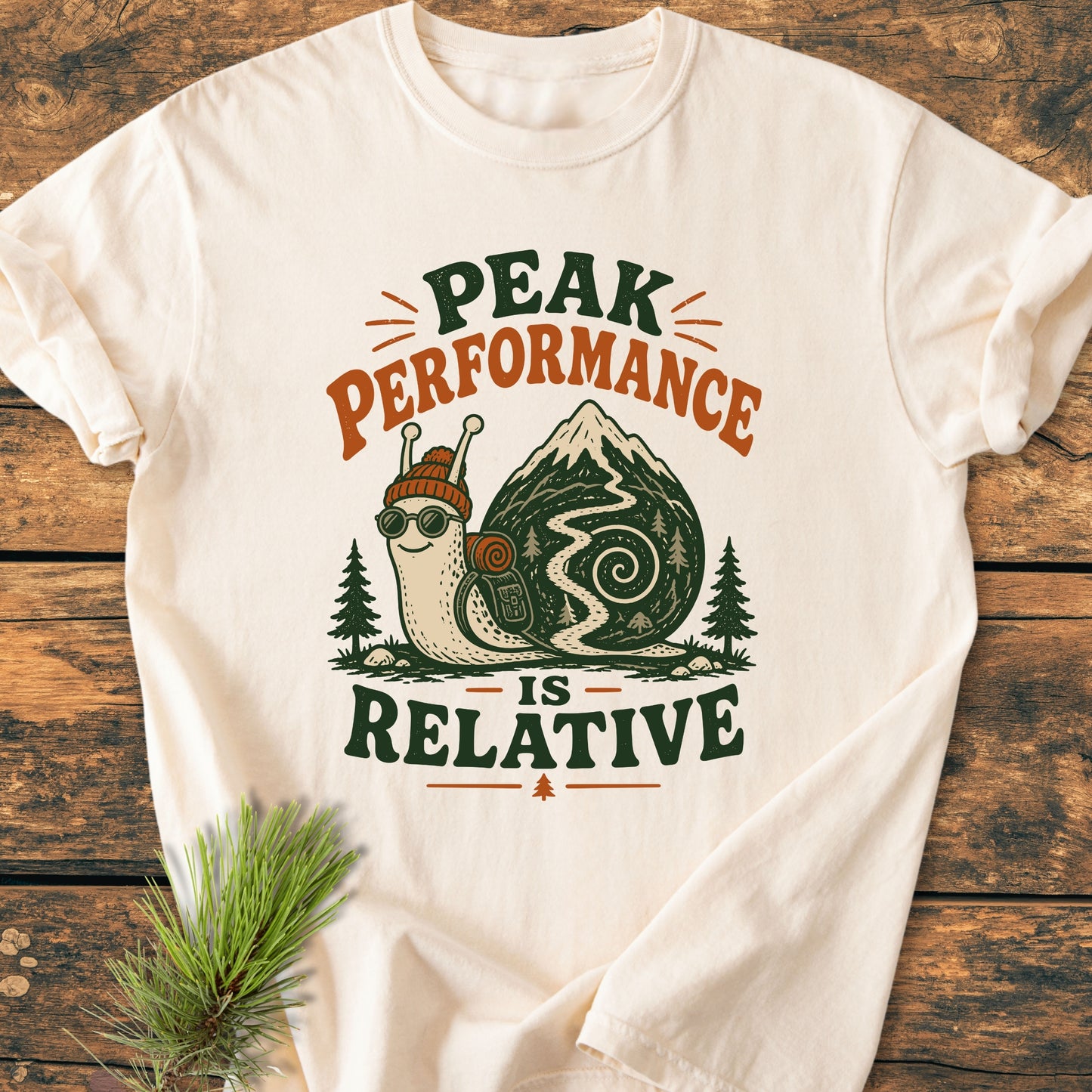 Off-white T-shirt with a vintage-style hiking snail print, slogan PEAK PERFORMANCE IS RELATIVE, laid on wood, with pine branches. Perfect for laid-back hikers.