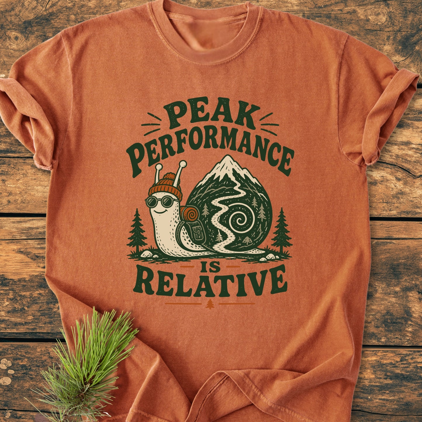 Peak Performance Is Relative - Vintage Wash Tee featuring a hiking snail graphic with mountain shell, laid flat on wood surface, complemented by pine sprigs, embodying a relaxed, outdoor adventure theme.