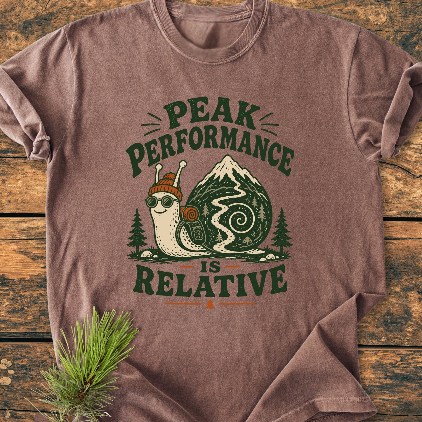 Heather-brown shirt featuring a cartoon snail with a knit cap and goggles, trail-shaped shell, and slogan “Peak Performance Is Relative” for laid-back adventurers. Perfect for slow-paced, nature-loving hikers.