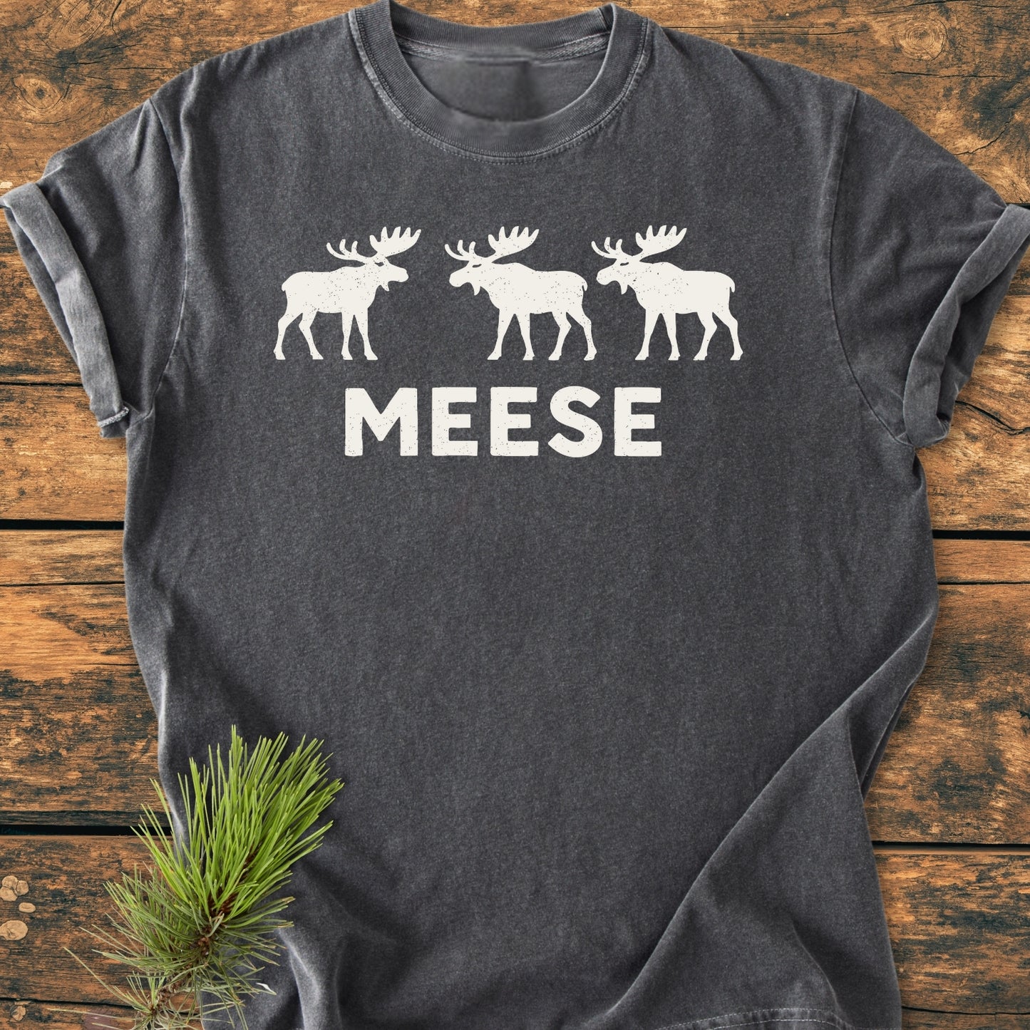 Charcoal-gray Meese - Vintage Wash Tee with white moose silhouettes and MEESE text, laid flat on wood. Pine branch decorates the corner, highlighting its outdoor theme and humorous design.