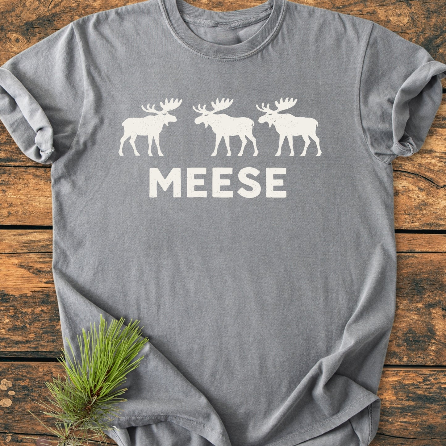 Gray Meese - Vintage Wash Tee with three white moose silhouettes and MEESE text, laid on wood surface with pine branch, embodying outdoor humor and relaxed unisex fit.