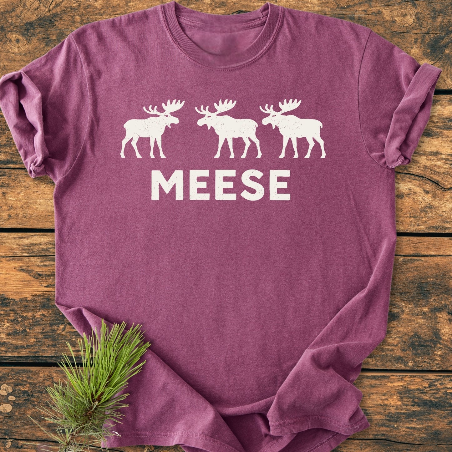 Heather-purple Meese - Vintage Wash Tee with white moose silhouettes and MEESE caption, laid flat on rustic wood beside a spruce sprig, showcasing its relaxed fit and screen-printed design.