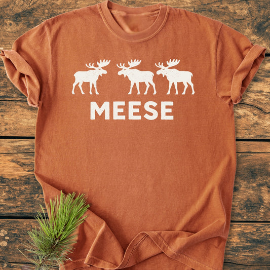 Rust-orange T-shirt titled Meese - Vintage Wash Tee with white moose silhouettes and MEESE text on the chest; pine sprig alongside, emphasizing outdoorsy humor and vintage style.