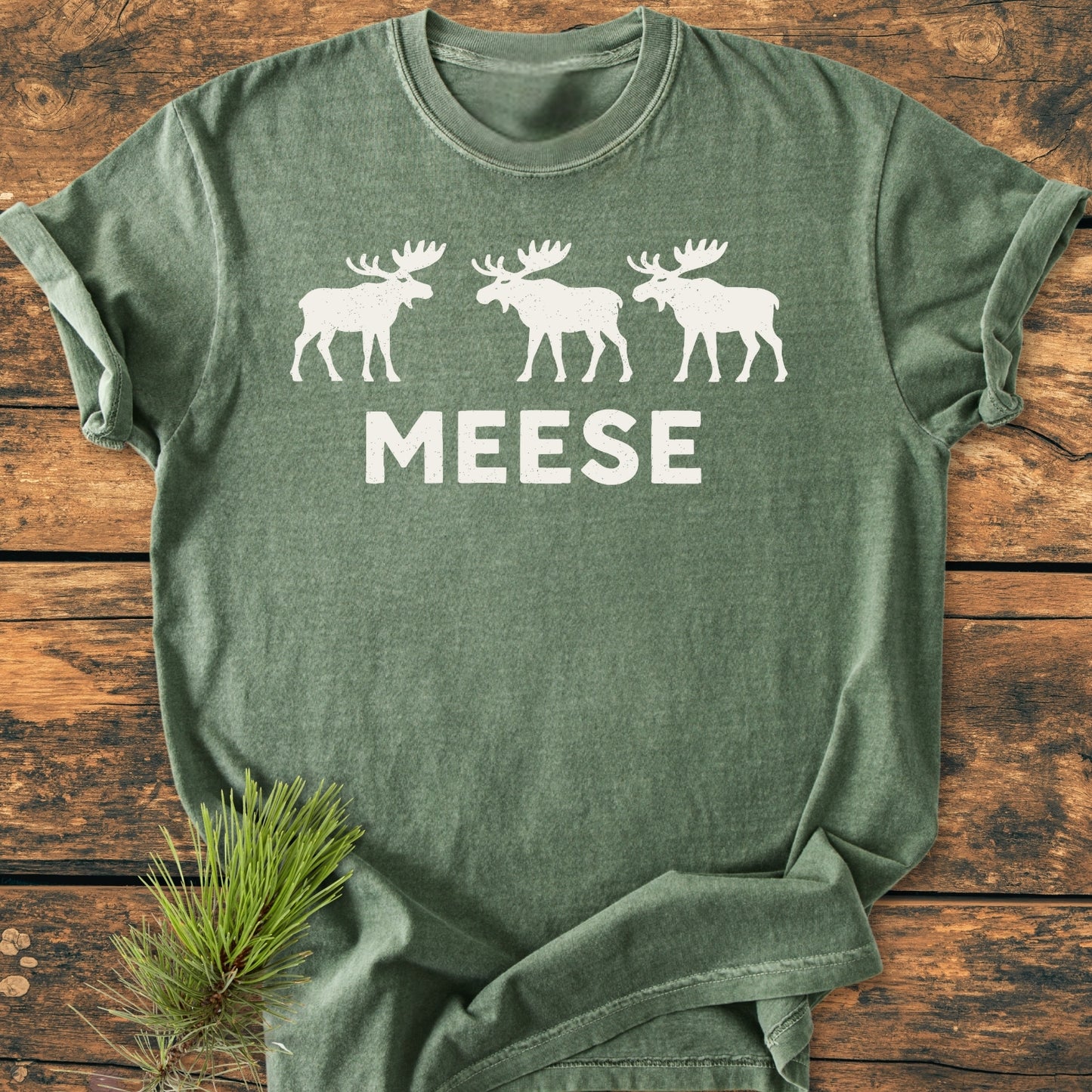 Heather-green Meese - Vintage Wash Tee with three white moose silhouettes and MEESE text, laid on wood with pine sprig. Ideal for moose lovers and outdoor humor enthusiasts.