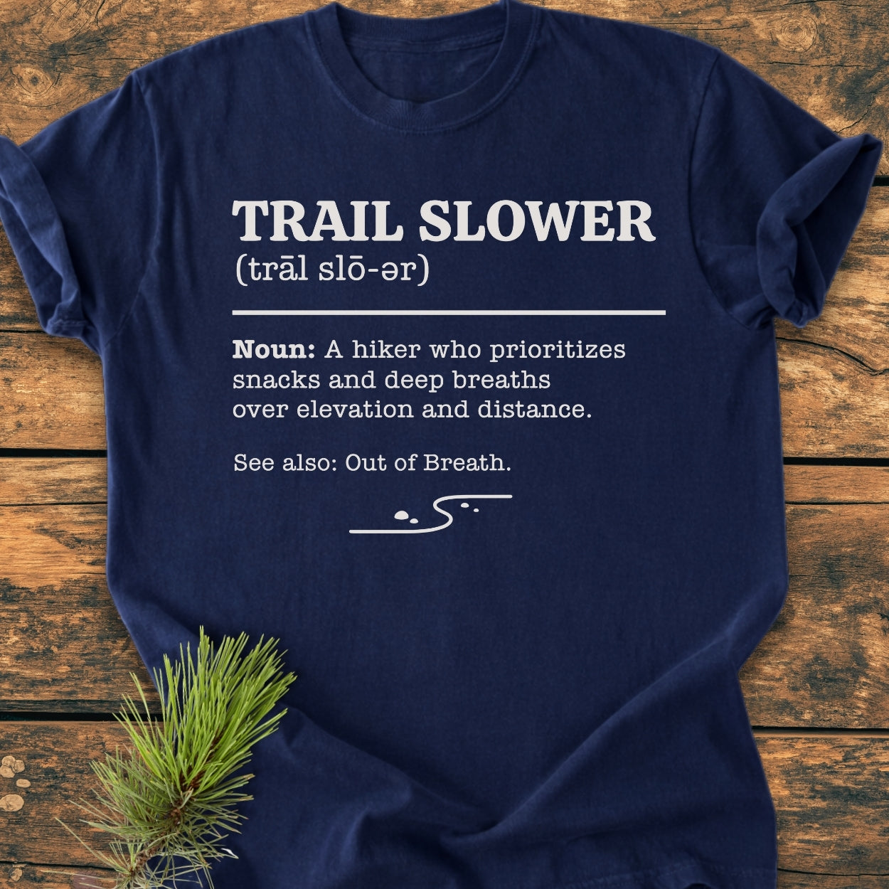 Navy-blue Trail Slower Dictionary - Vintage Wash Tee featuring a humorous white mock-dictionary entry and a stylized trail graphic, laid out on a wooden surface, embodying a relaxed hiking lifestyle.