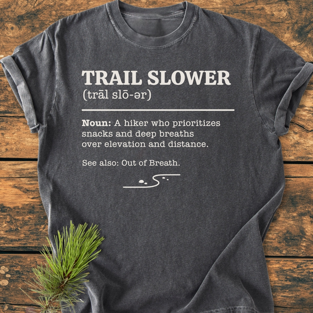 Charcoal-gray Trail Slower Dictionary - Vintage Wash Tee with humorous hiking definition in white text and winding trail design, laid on wood with a pine branch accentuating its outdoor vibe.