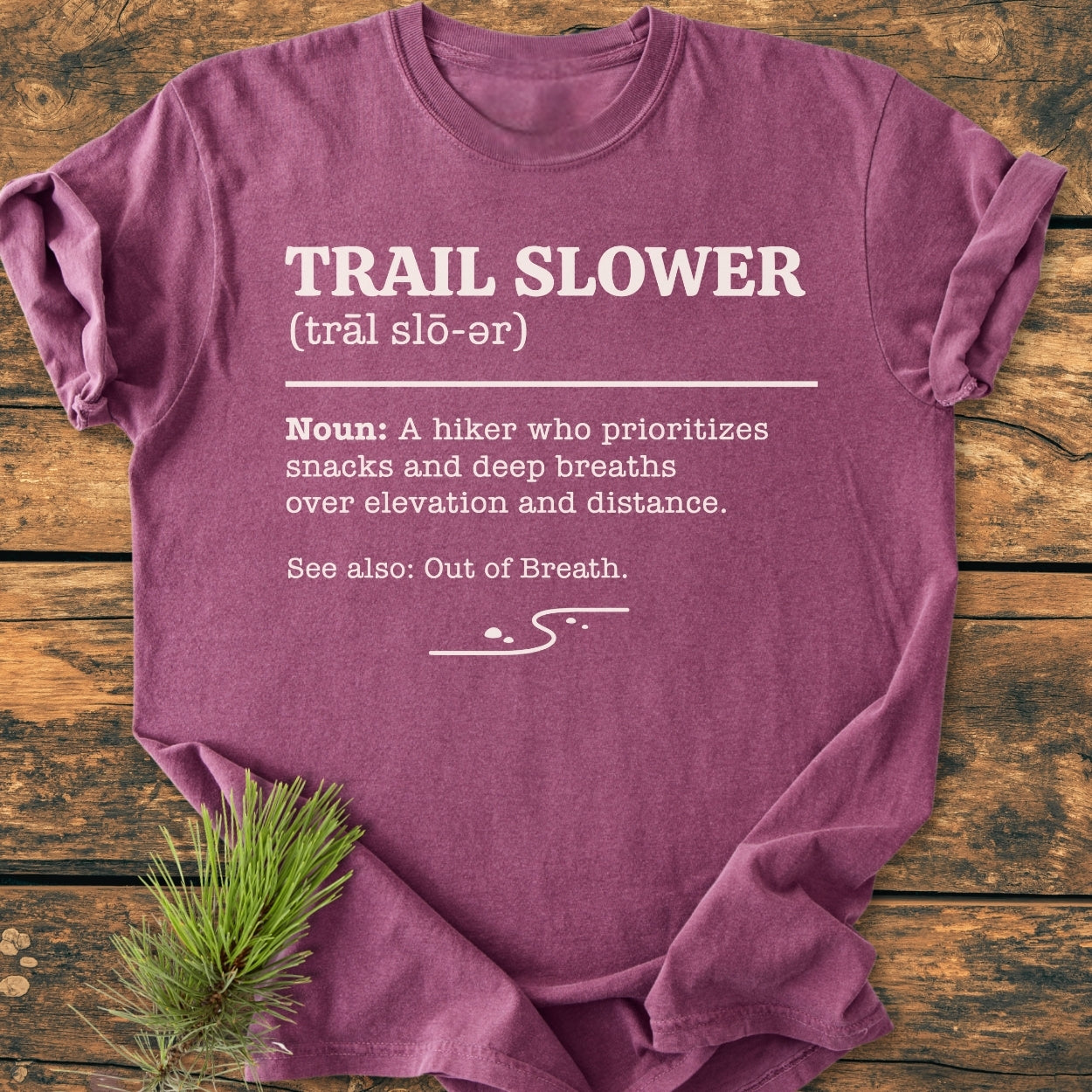 The maroon Trail Slower Dictionary - Vintage Wash Tee features white dictionary-style text and a trail graphic, laid on rustic wood with a pine branch beside it, highlighting its relaxed, humorous hiking theme.