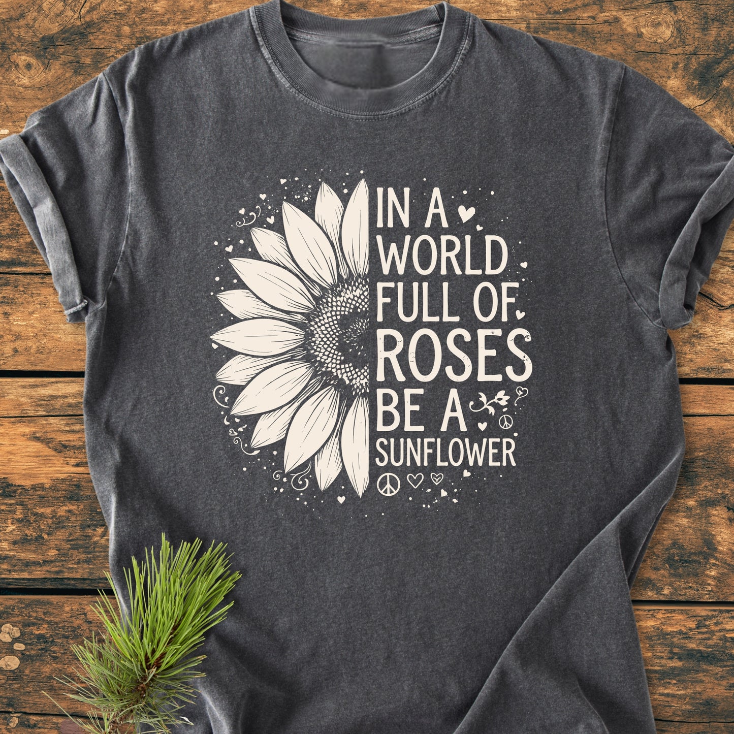 Be A Sunflower - Vintage Wash Tee: Flat-lay of a dark T-shirt featuring a half-sunflower illustration and the slogan, laid on a wooden surface with a pine sprig, showcasing a relaxed, unisex fit.