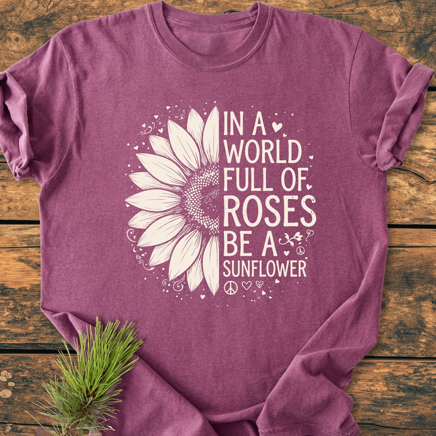 Be A Sunflower - Vintage Wash Tee, featuring half a sunflower and slogan in white, displayed on a wooden surface with an evergreen sprig, highlighting its nature-inspired, feel-good design.