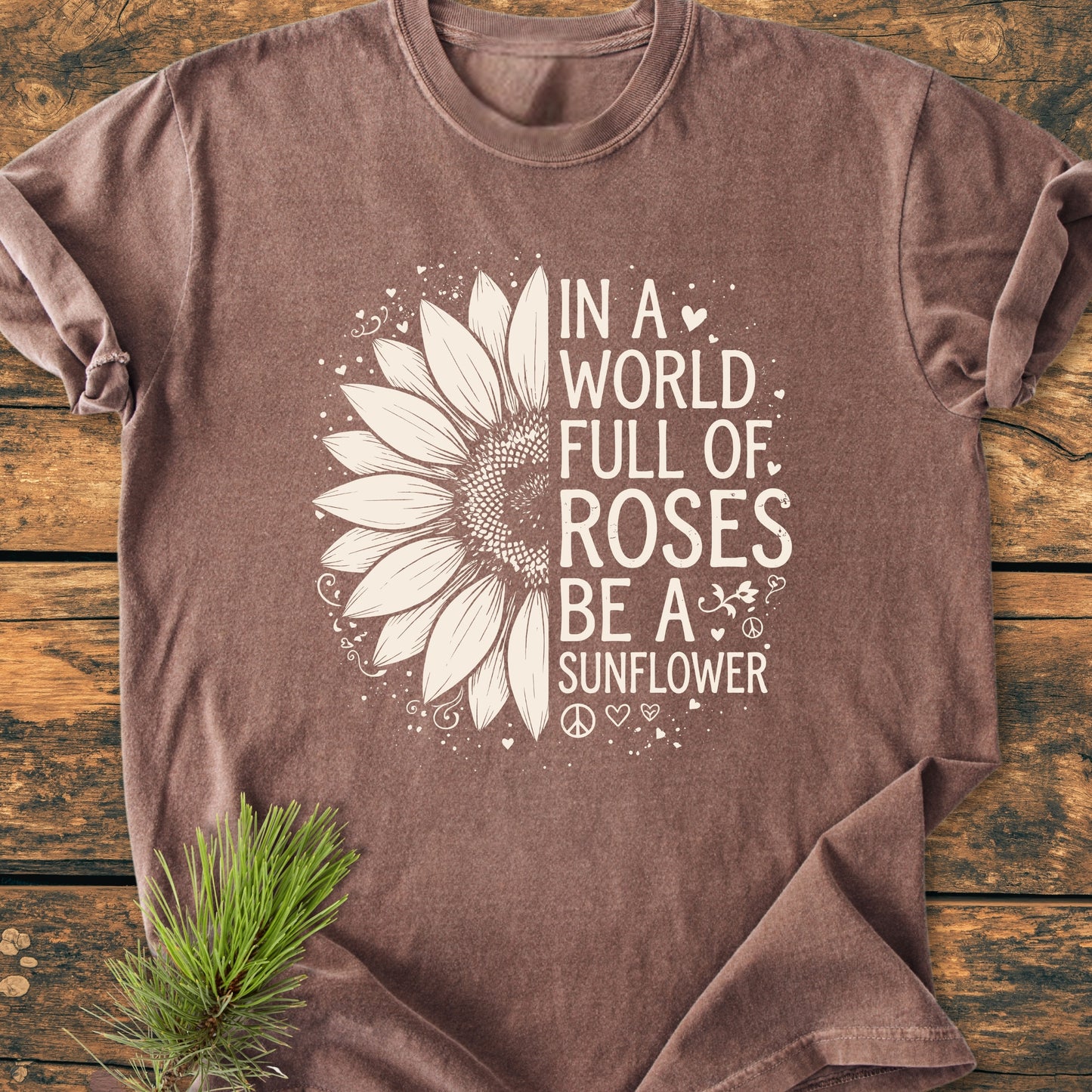 Be A Sunflower - Vintage Wash Tee features a sunflower graphic and inspirational text, laid flat on wood, with pine needles accent. Perfect for those embracing warmth and individuality.