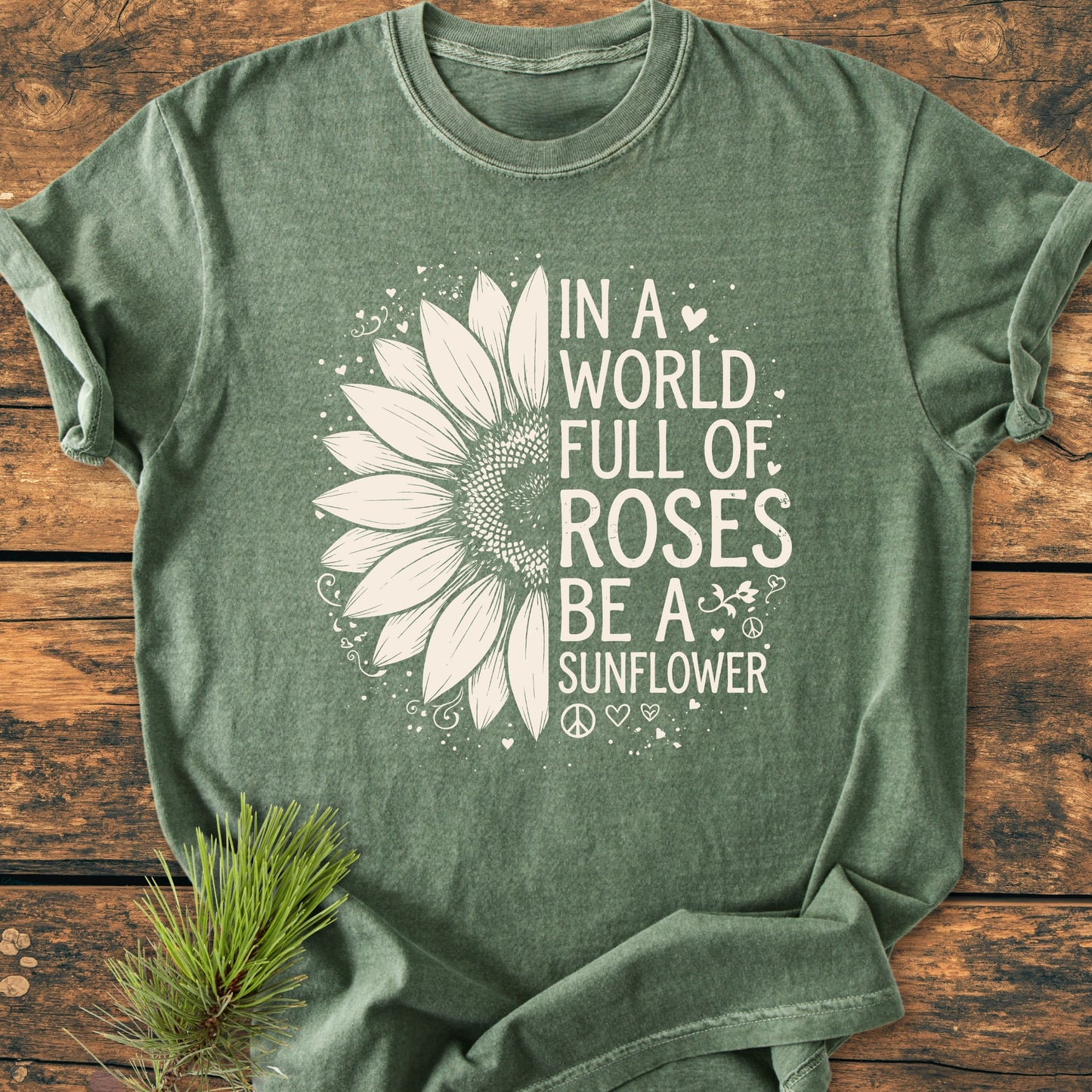 Be A Sunflower - Vintage Wash Tee featuring a white sunflower graphic and slogan on a green heather T-shirt, laid on wooden surface with a pine branch beside it.