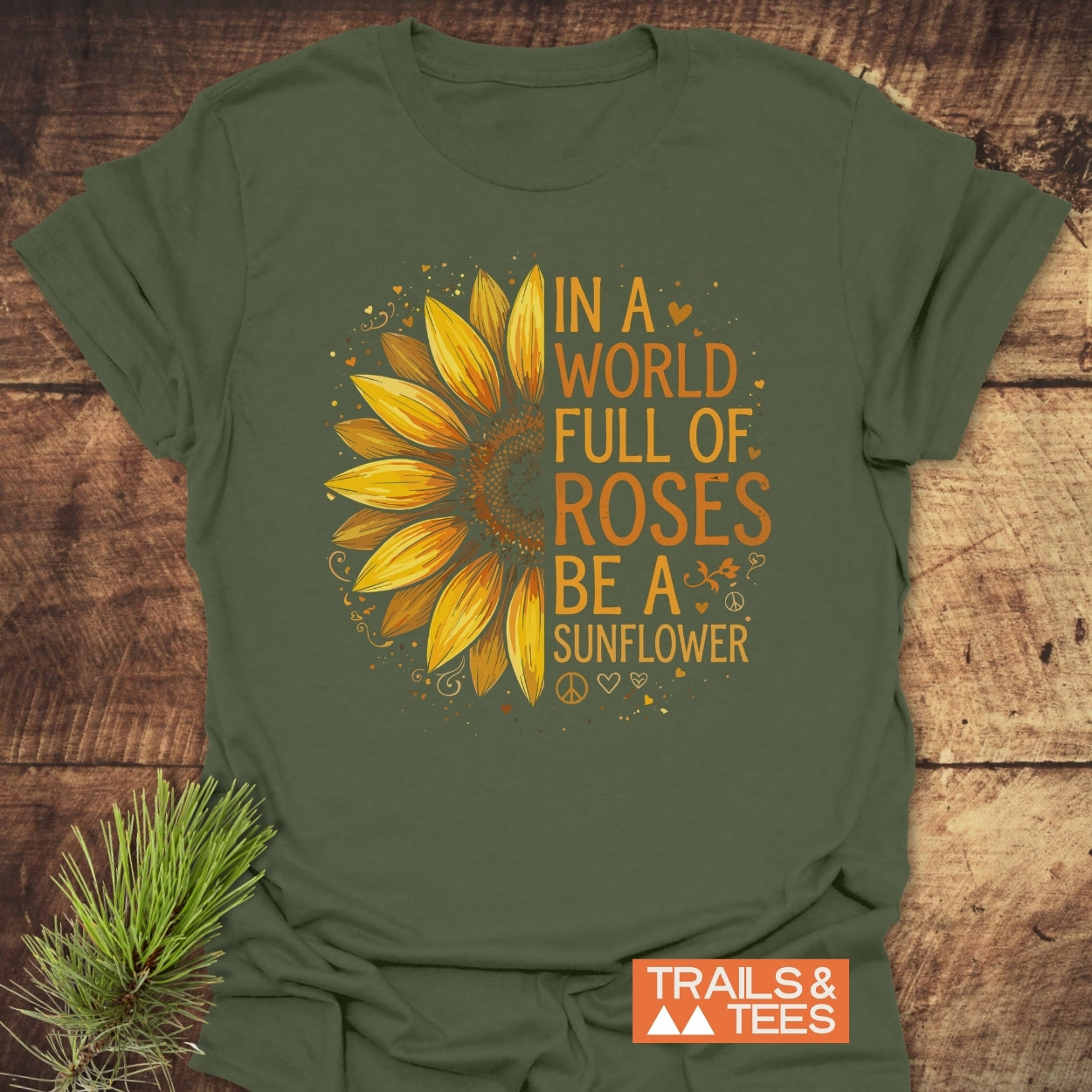 Be A Sunflower Softstyle T-Shirt: Olive-green tee with half-yellow sunflower and uplifting text. Features bold orange-yellow slogan and Trails & Tees logo, laid on wood with pine sprig.