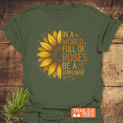 Be A Sunflower Softstyle T-Shirt: Olive-green tee with half-yellow sunflower and uplifting text. Features bold orange-yellow slogan and Trails & Tees logo, laid on wood with pine sprig.