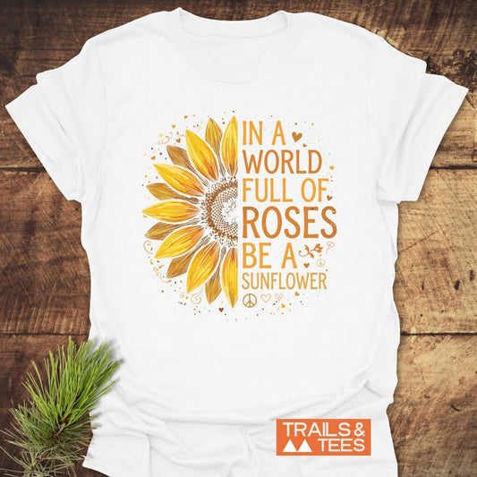 Be A Sunflower Softstyle T-Shirt, featuring a sunflower graphic with the slogan In a world full of roses be a sunflower, surrounded by tiny hearts and peace signs, on a wooden surface.