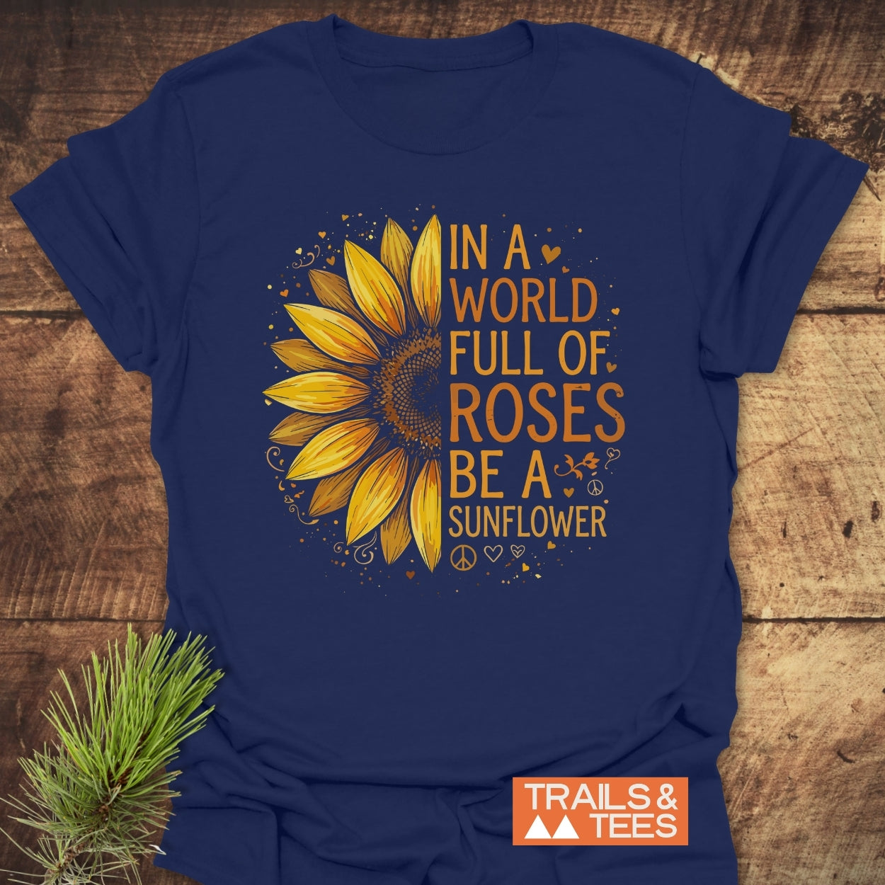 Be A Sunflower Softstyle T-Shirt featuring a stylized half-sunflower with hearts, swirls, and peace signs, slogan beside it. Laid flat, navy-blue, with Trails & Tees logo in the corner.
