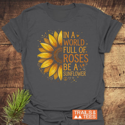 Be A Sunflower Softstyle T-Shirt on a wooden surface, featuring a half-sunflower graphic with hearts and peace symbols, and the phrase “In a world full of roses be a sunflower.”