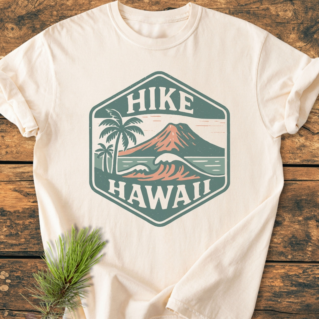 Hike Hawaii - Vintage Wash Tee with a hexagon crest featuring palm trees, waves, and a volcano, laid on wood with pine needles nearby, embodying island adventure and nature-inspired design.