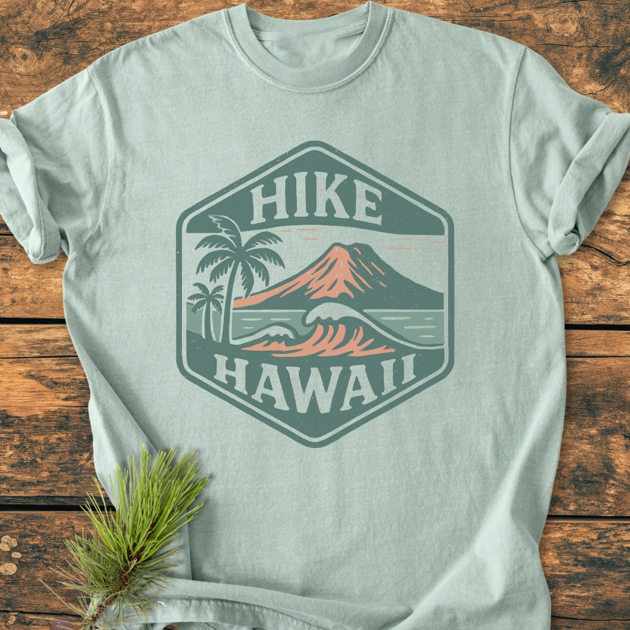 Pale-green Hike Hawaii - Vintage Wash Tee featuring a hexagonal badge with a stylized volcano, palm trees, and ocean waves, laid flat on rustic wood with a sprig of pine nearby.