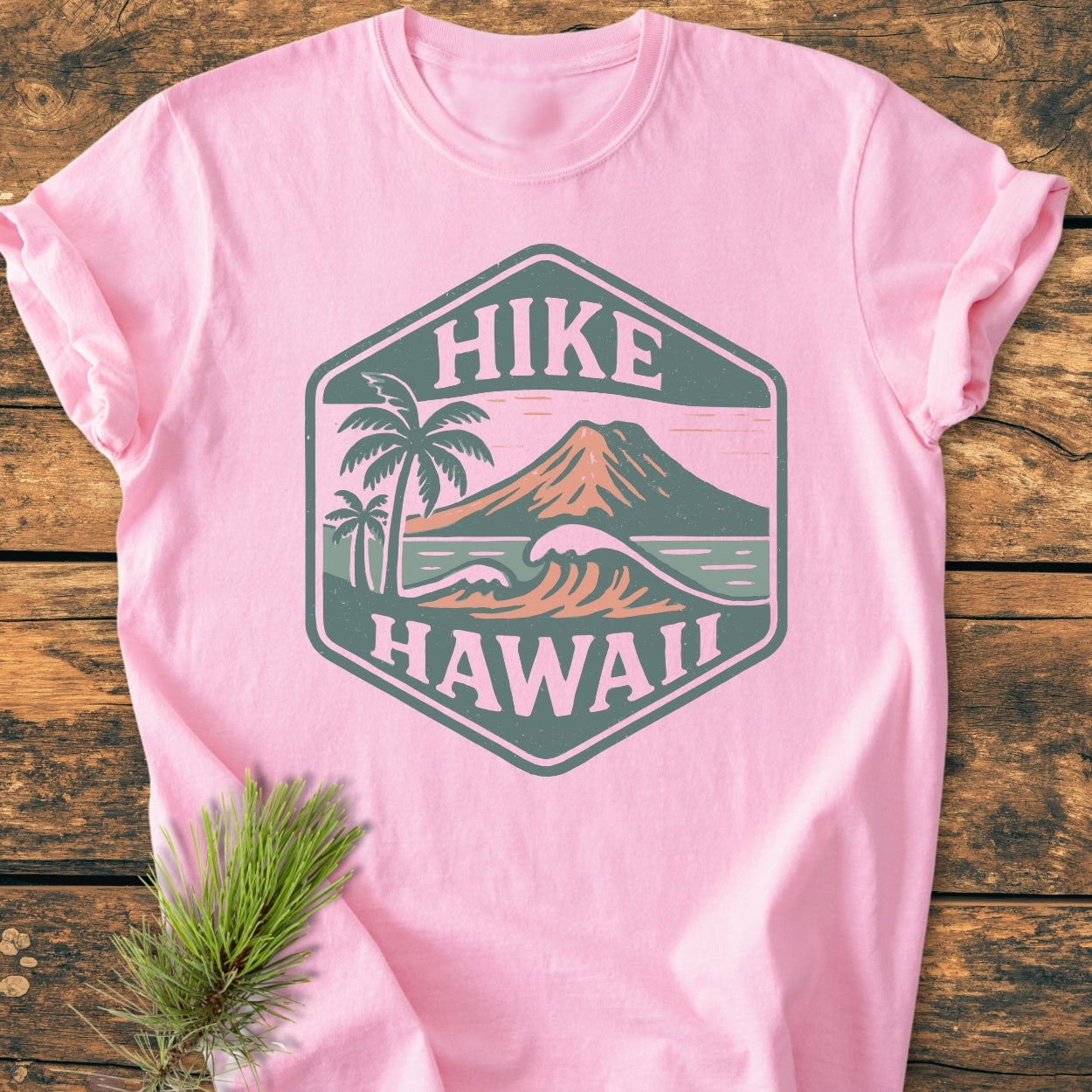 Hike Hawaii - Vintage Wash Tee: Light-pink cotton shirt with a HIKE HAWAII hexagon badge featuring a volcanic mountain, palm trees, and ocean waves, laid on a wooden surface with a pine sprig beside it.