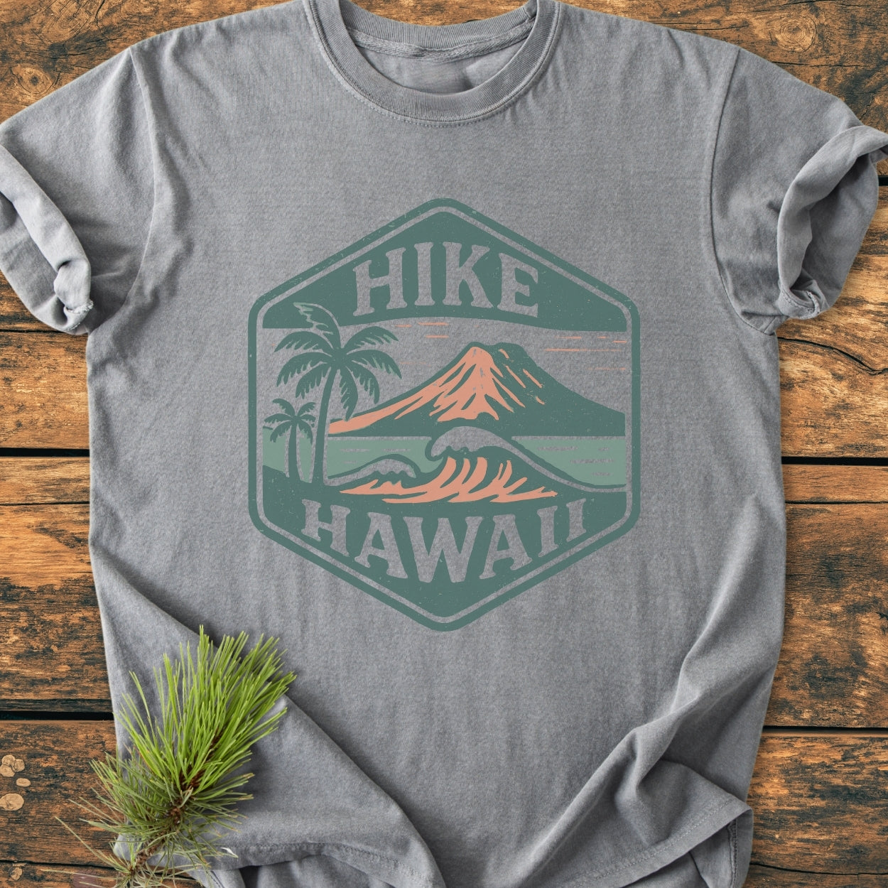 Heather-gray T-shirt named Hike Hawaii - Vintage Wash Tee, featuring a hexagonal badge with palm trees, volcanic mountain, and ocean waves. Rustic text reads “HIKE” and “HAWAII.” Pine sprig accents corner.