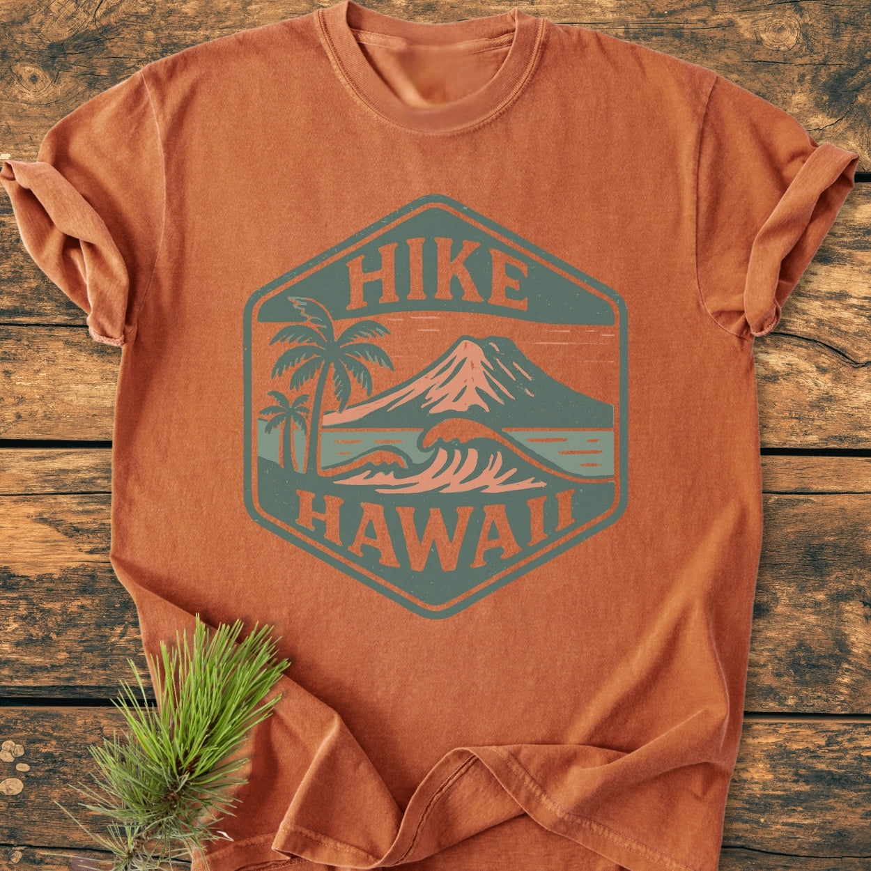 Hike Hawaii - Vintage Wash Tee featuring a hexagonal badge with palm trees, wave, and volcano, ideal for outdoor enthusiasts and island adventurers, laid on a wooden surface with a pine sprig.