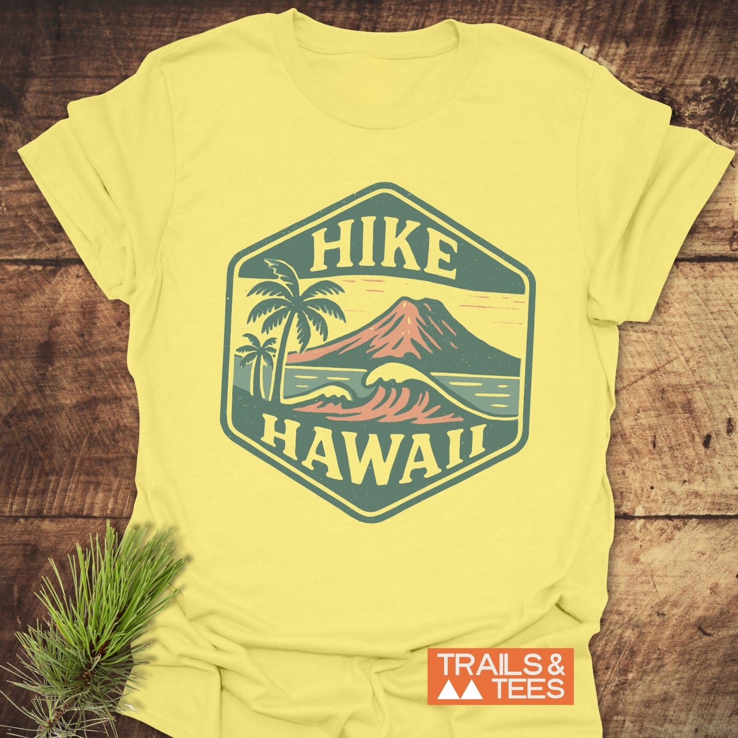 Hike Hawaii - Softstyle T-Shirt featuring a vintage tropical scene with a volcano, waves, and palm trees; TRAILS & TEES logo present, laid on a wooden surface with pine sprigs.